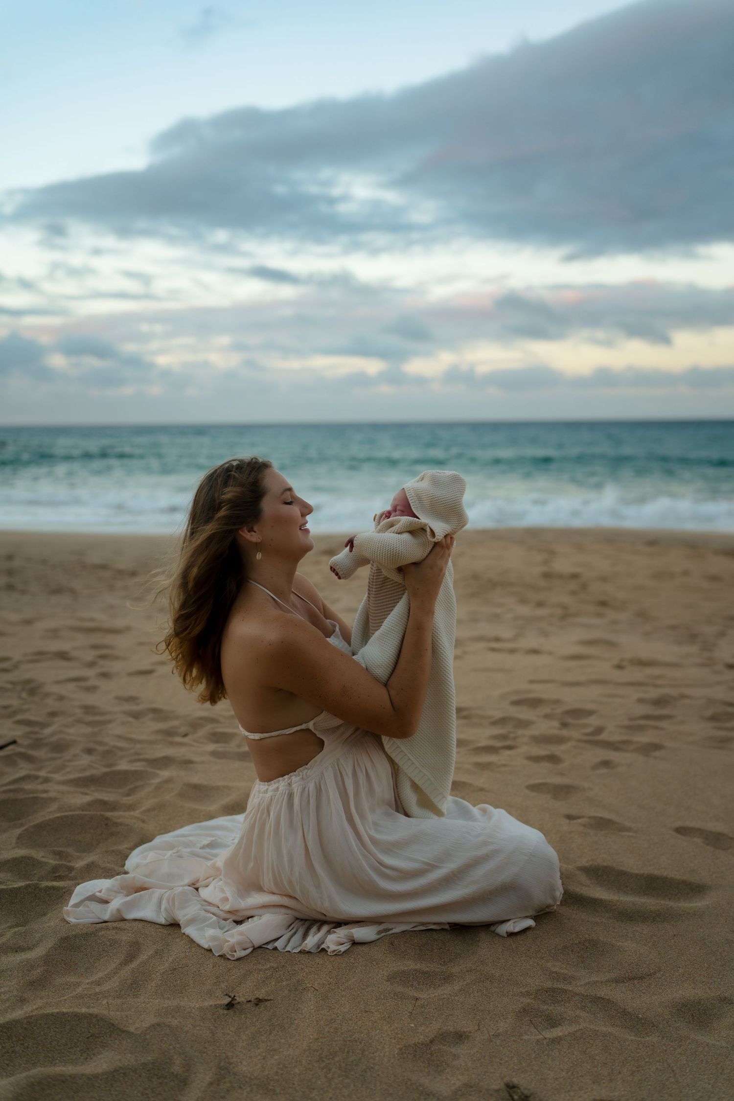 A mom in a white dress sits on a sandy beach at sunset holding her baby in a white blanket.
