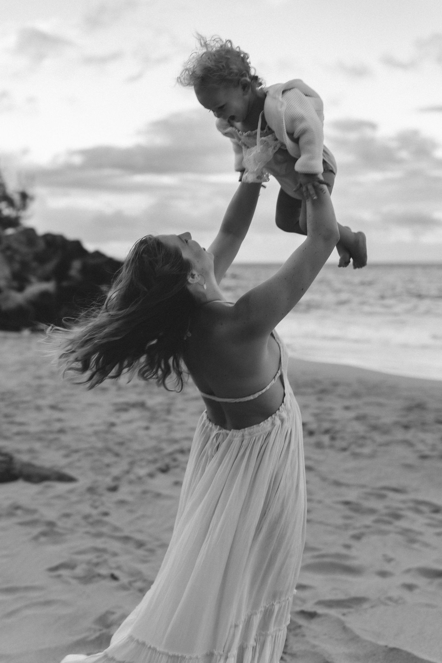 Woman in flowing white dress plays joyfully with baby on beach at sunset.