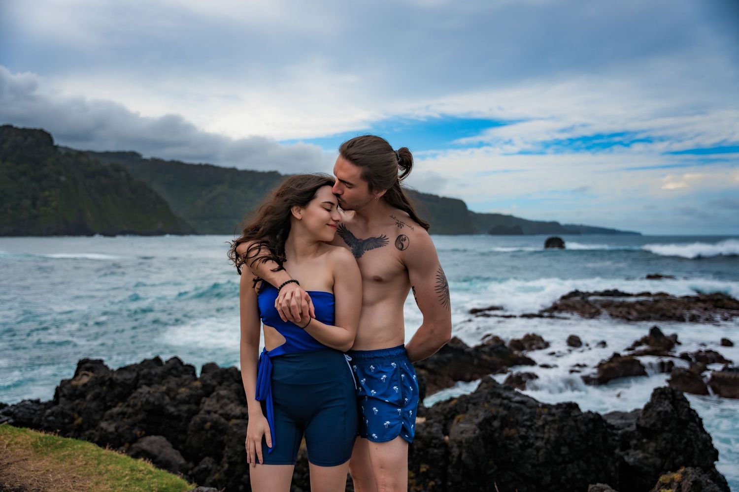 A couple shares intimate moments along a rocky coastline with ocean waves and blue skies.
