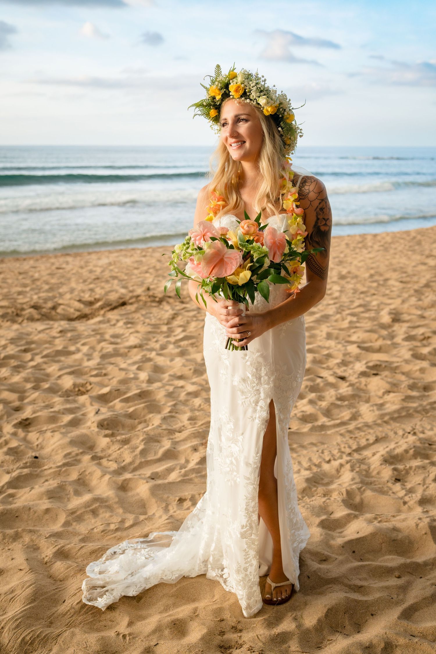 A bride in a lace dress with floral crown holds a colorful bouquet on a tropical beach.