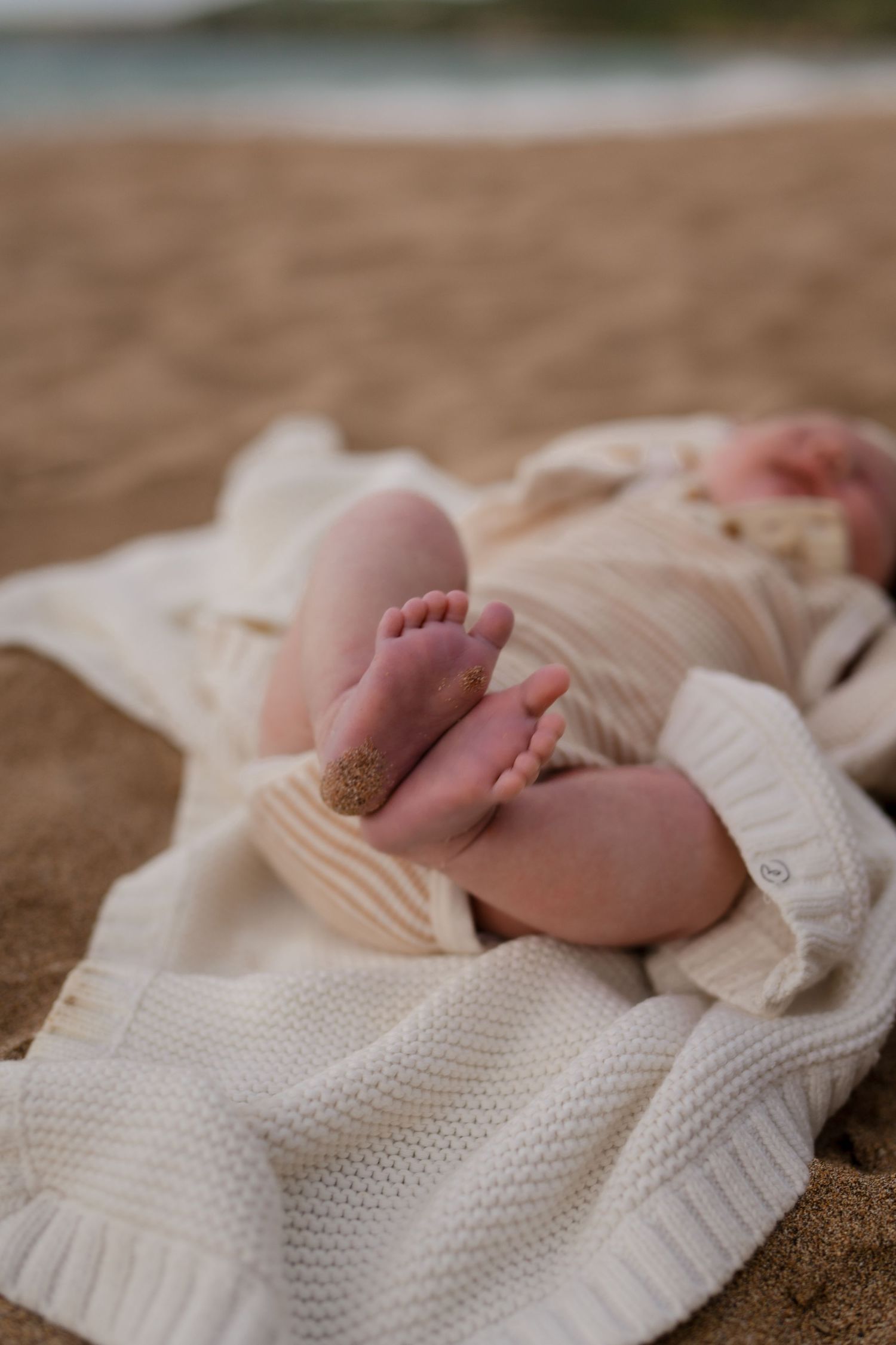 Tiny baby feet resting on a cozy white knitted blanket against a sandy beach background.