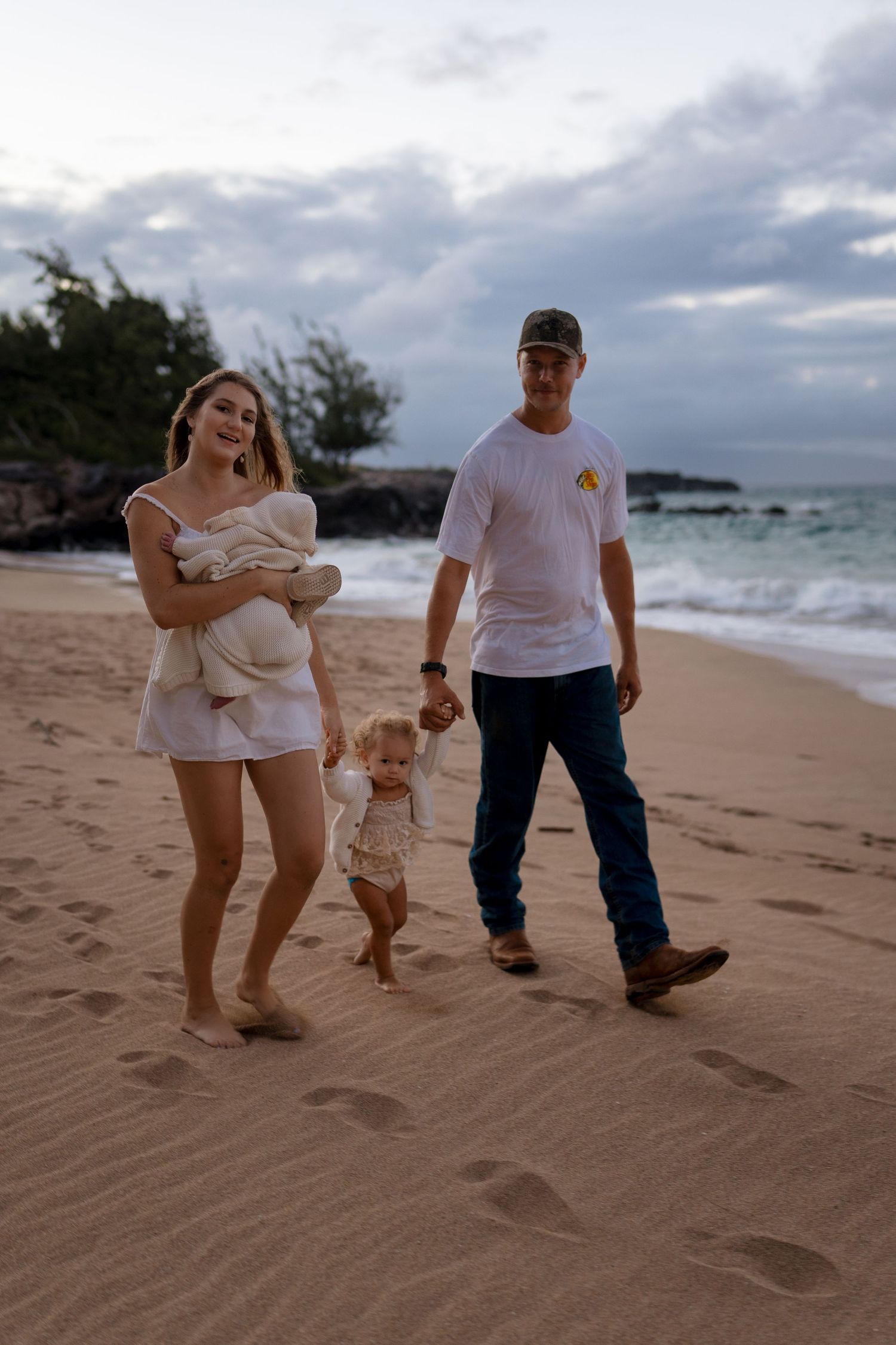 A family walks together on a sandy beach at sunset with cloudy skies and ocean waves in the background.