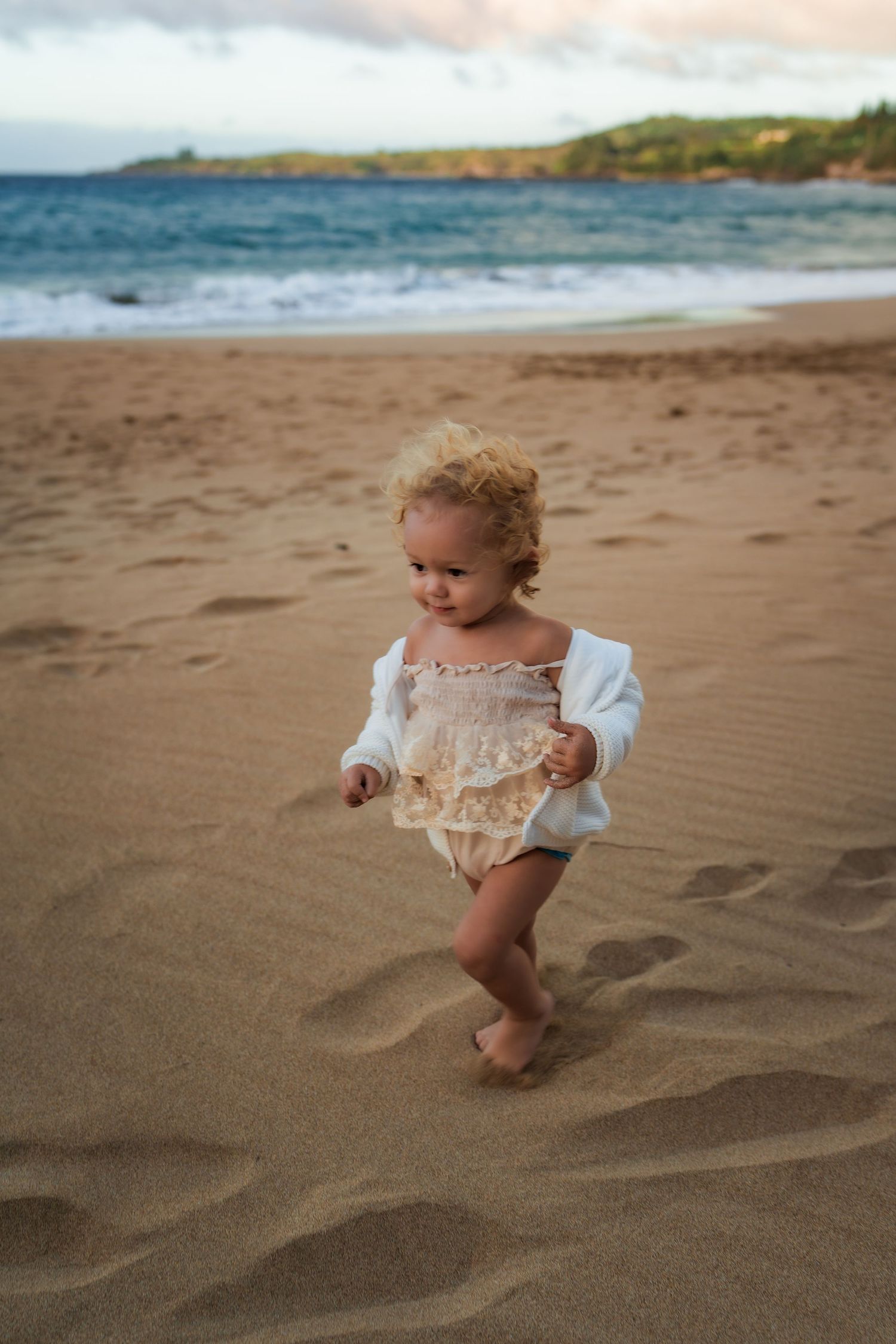 A toddler in a white outfit plays in the sand at the beach during golden hour.