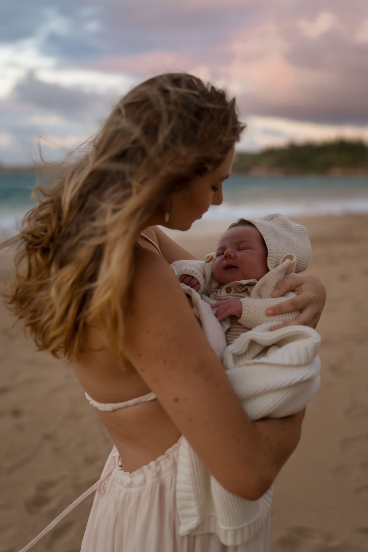 A tender moment between mom and newborn baby on a beach at sunset.