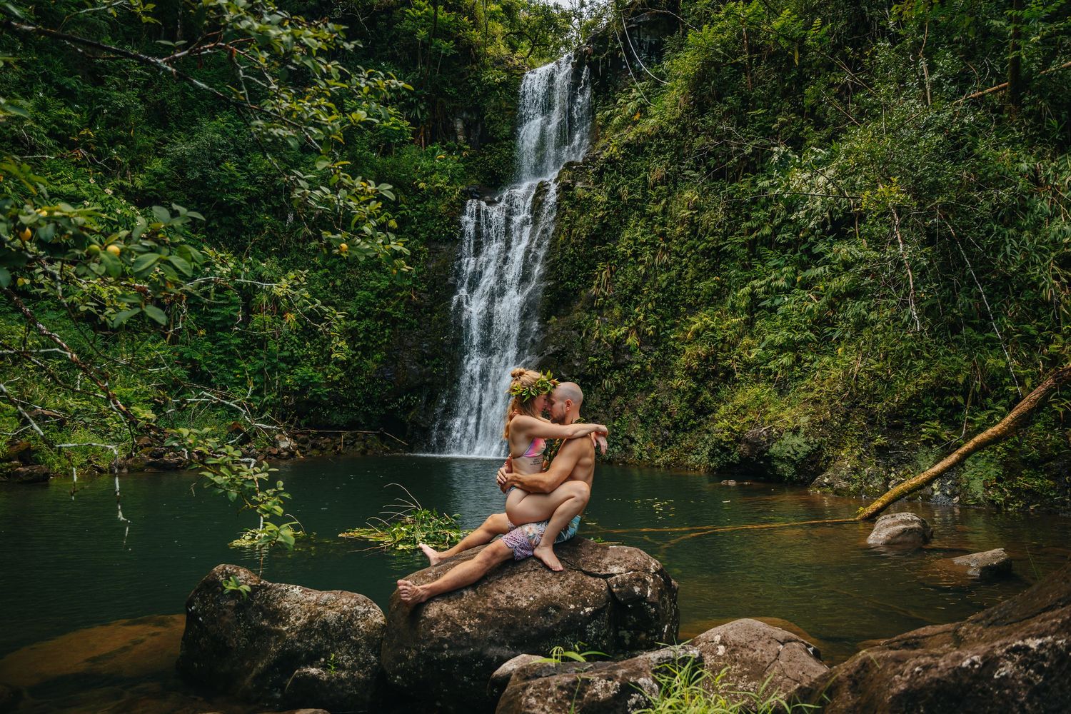Couple relaxing on large rocks near a scenic waterfall surrounded by tropical foliage.