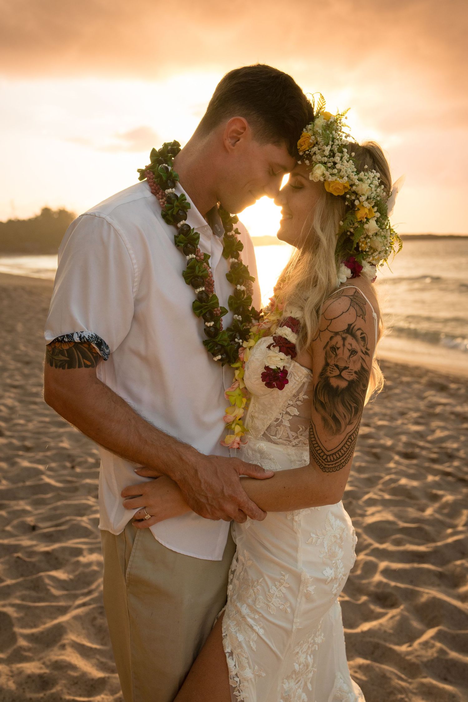 A tender moment between newlyweds wearing leis against a golden sunset on the beach.