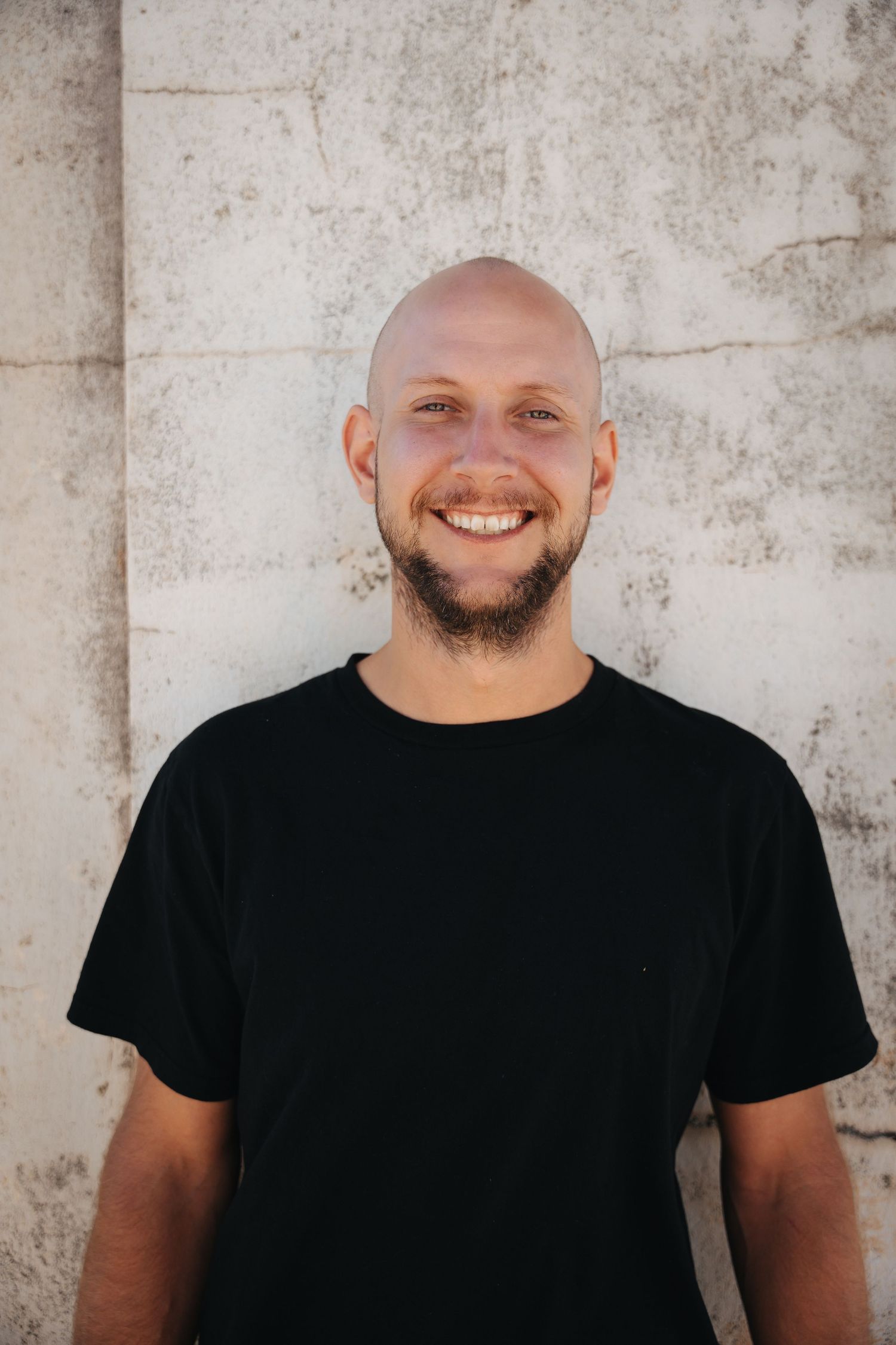 A person in a black t-shirt smiles warmly against a textured concrete wall background.