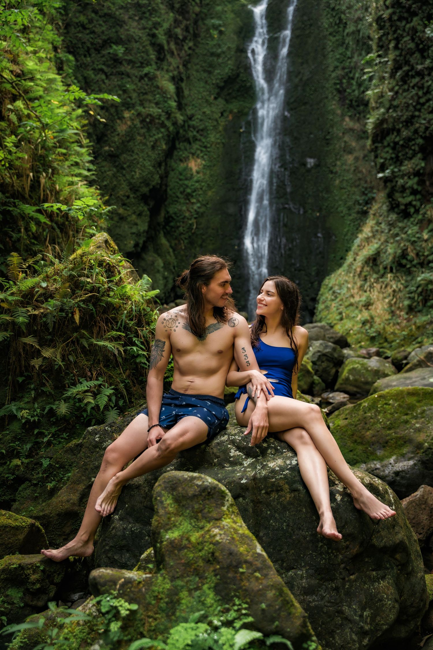 Two people sit on rocky outcrop with waterfall and lush greenery in background.