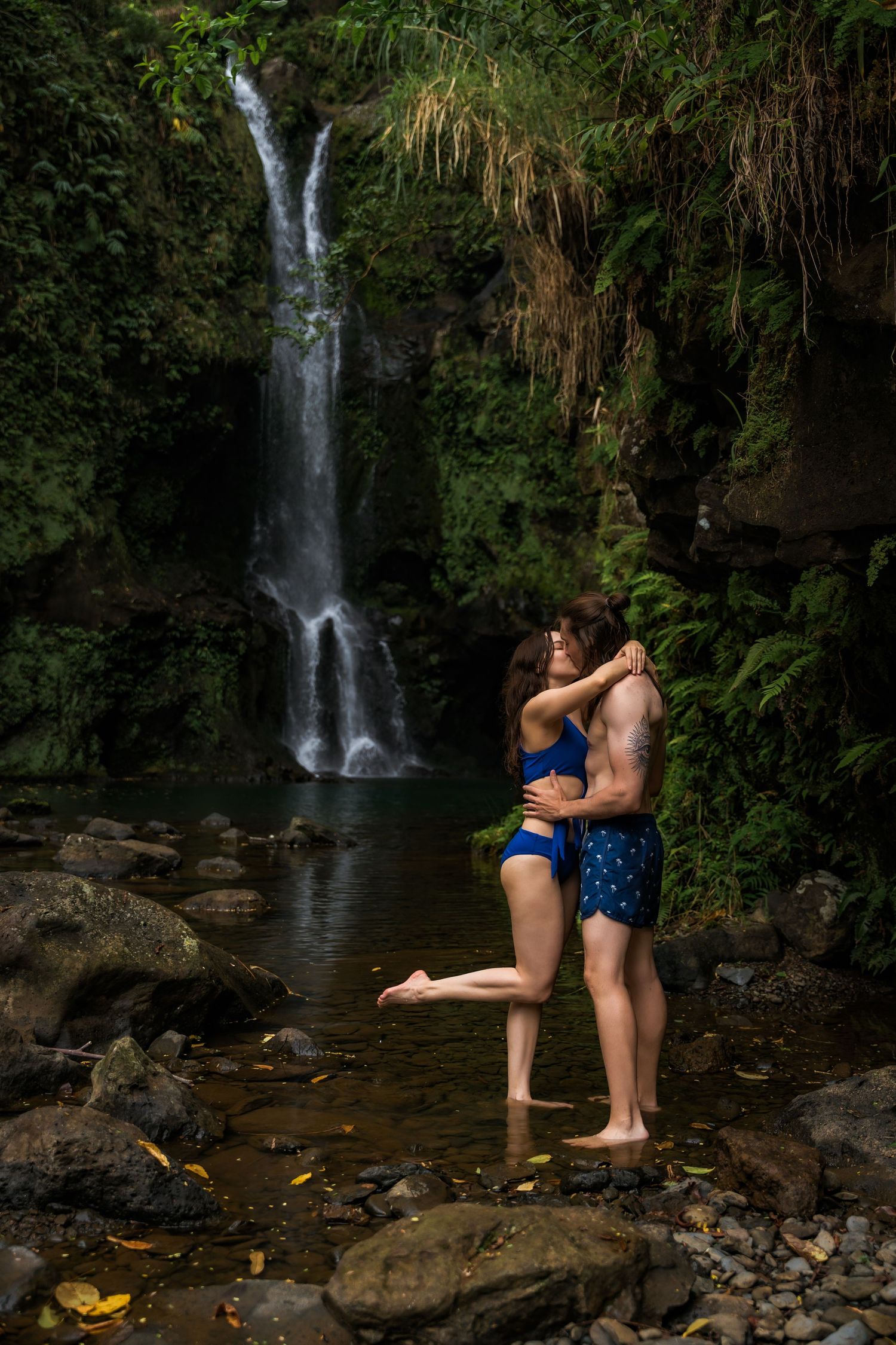 A romantic scene unfolds by a cascading waterfall in a lush tropical forest setting.
