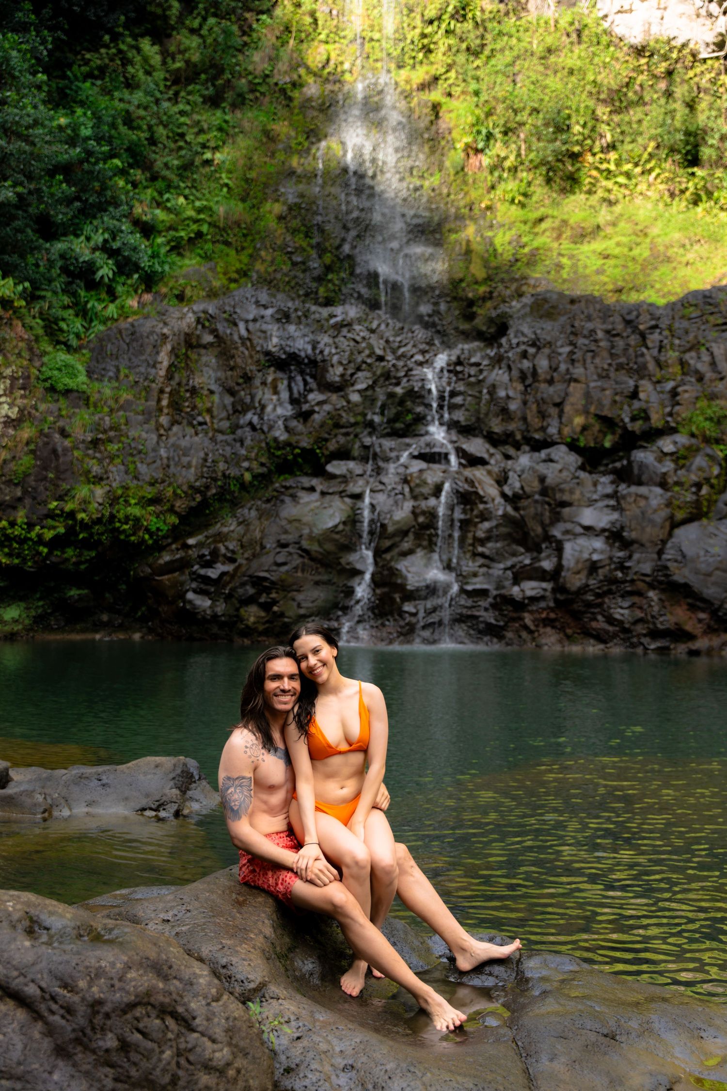 A couple relaxes on rocks beside a serene waterfall pool in a tropical paradise.