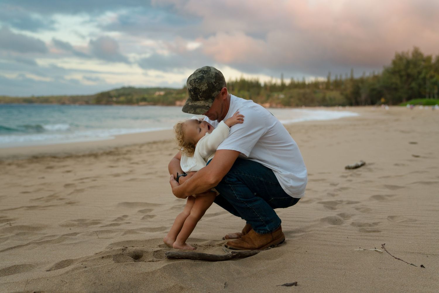 Candid beach scene with dad and daughter sitting on sand during sunset with ocean waves in background.