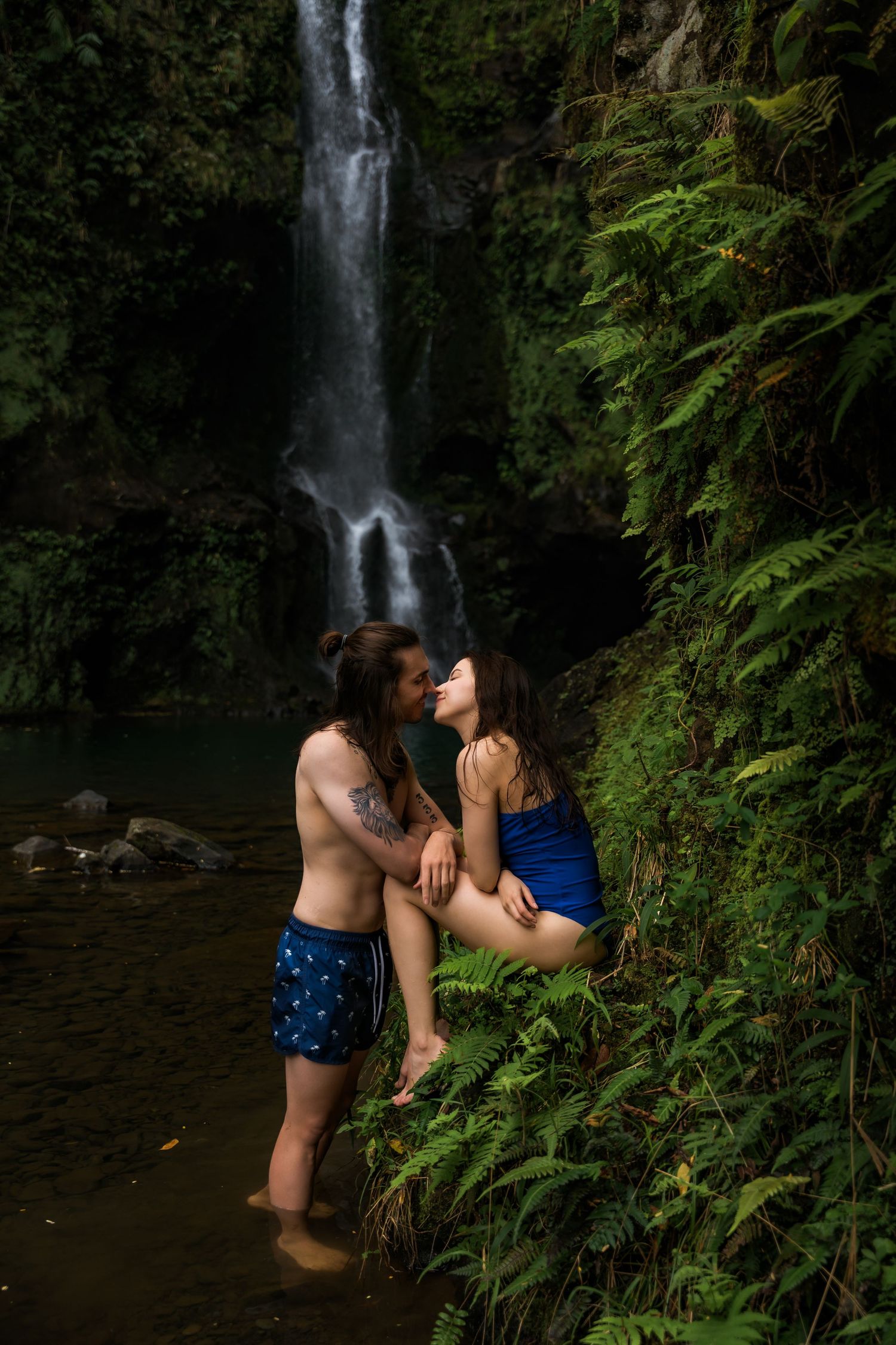 Romantic moment captured by a waterfall with two people standing in shallow water among ferns.