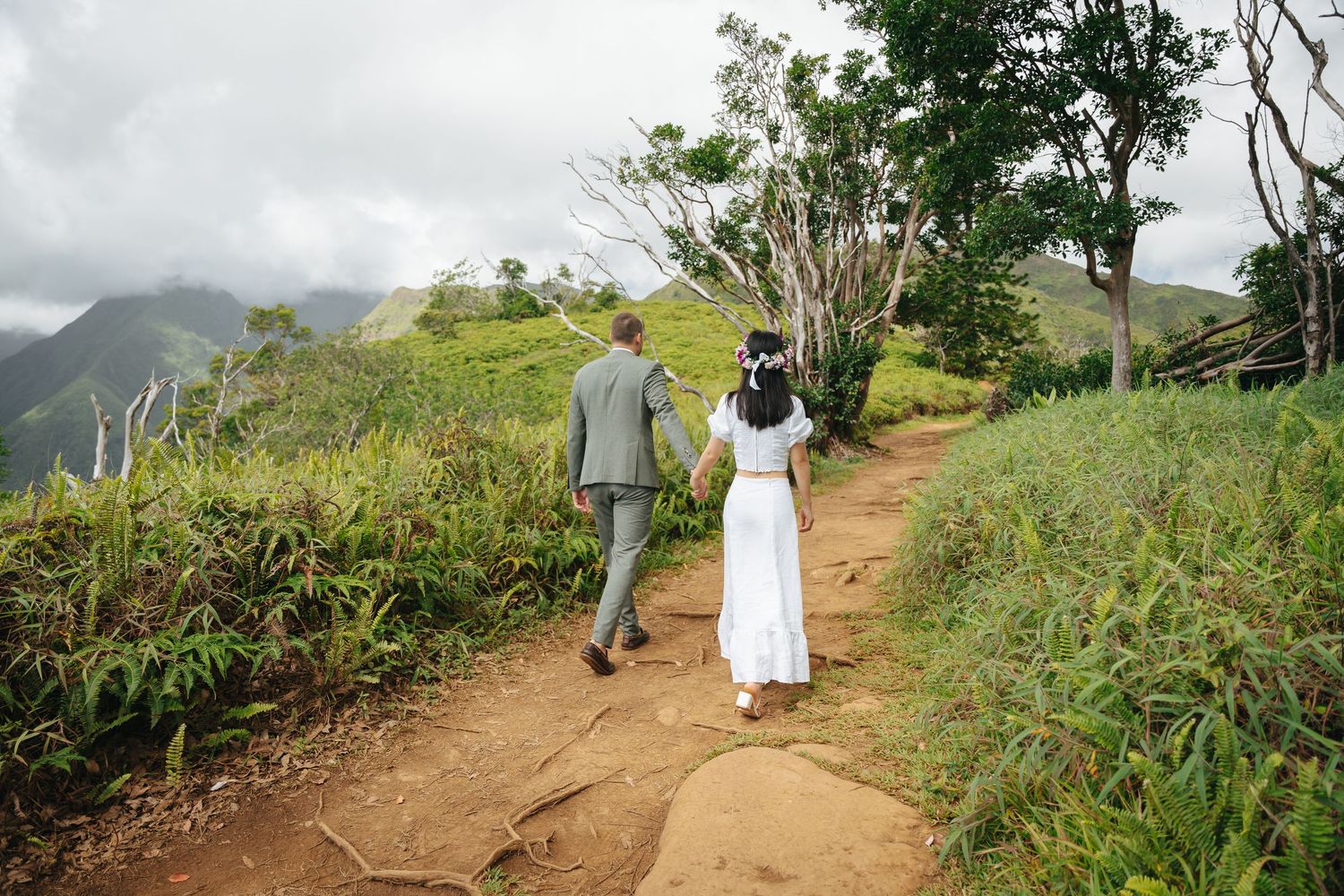 A scenic hiking trail winds through lush Hawaiian mountains with misty peaks in the distance.