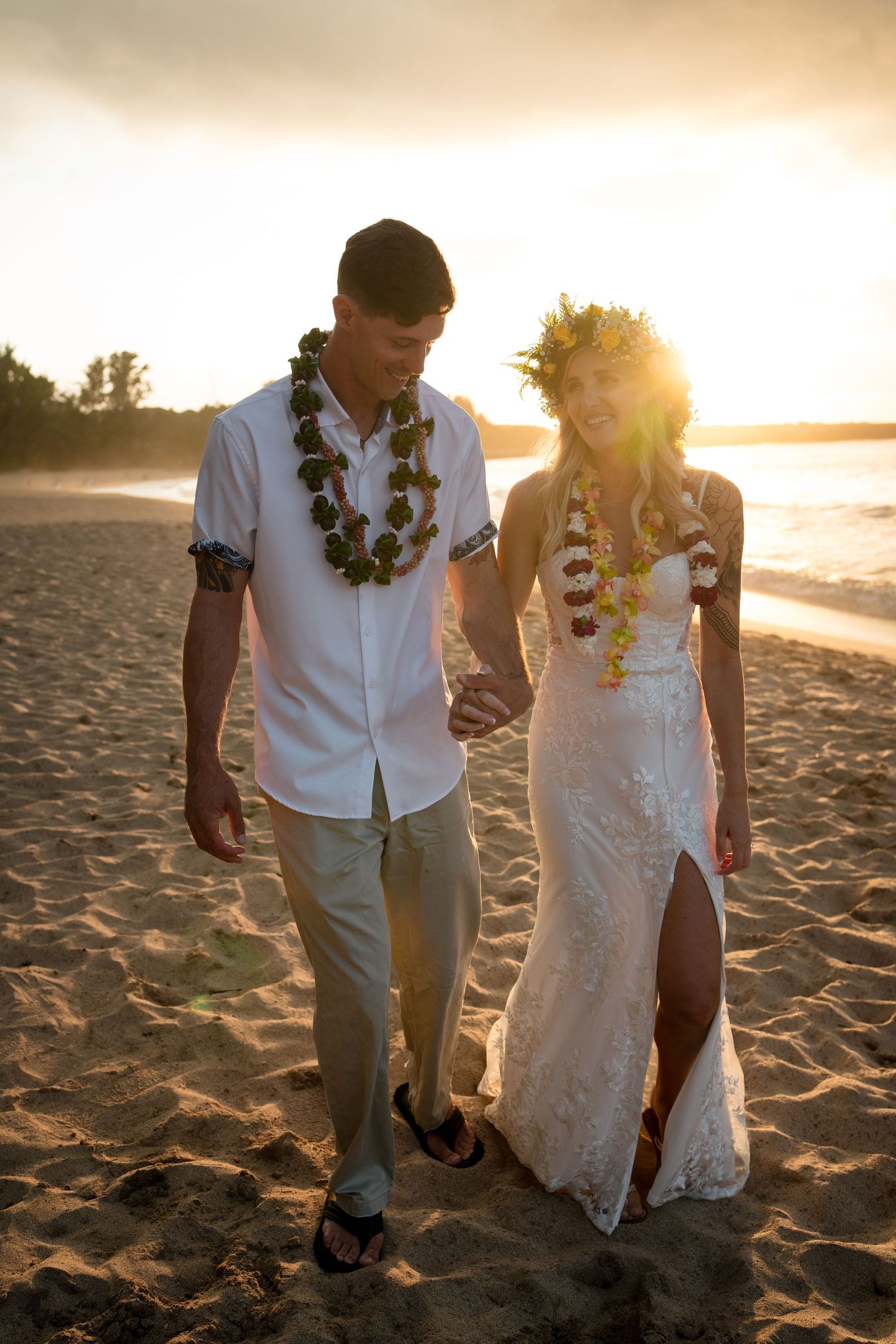 A couple in wedding attire walks along a beach at sunset with tropical leis around their necks.