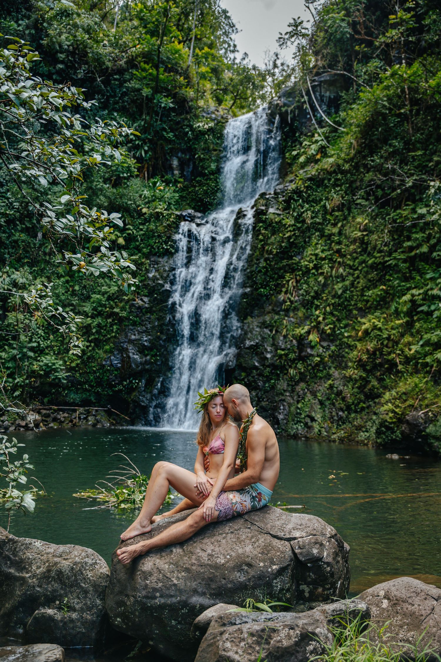 Romantic couple sitting on rocks by a cascading waterfall in a lush tropical setting.