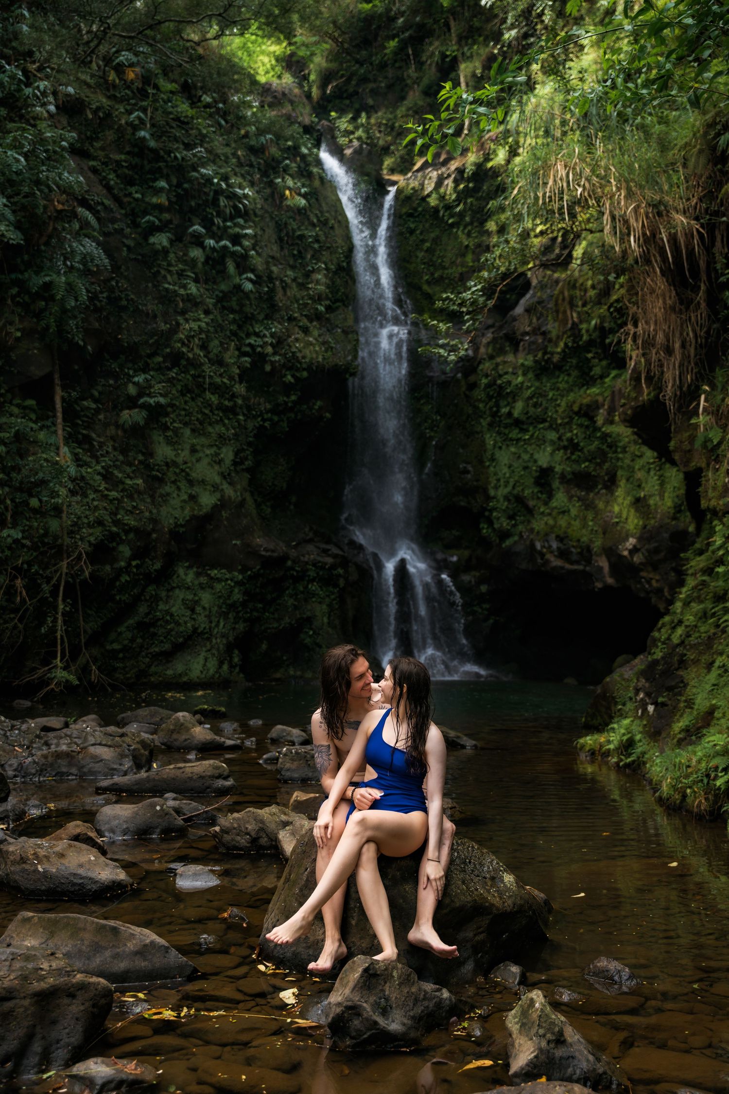 Two people share a tender moment on mossy rocks by a waterfall in a tropical setting.