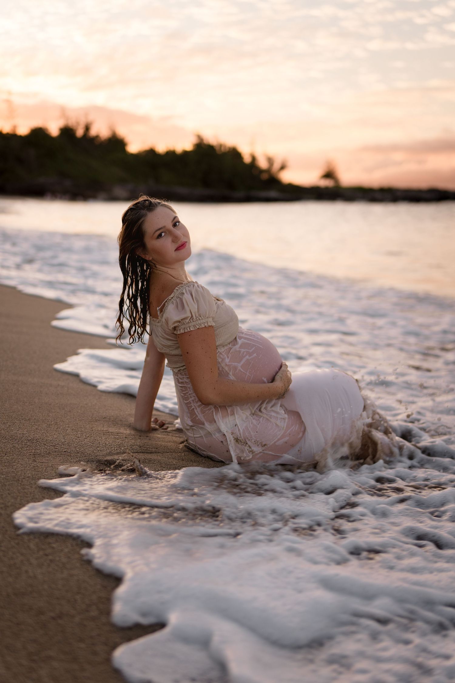 A maternity photo of someone sitting in beach waves during golden hour.