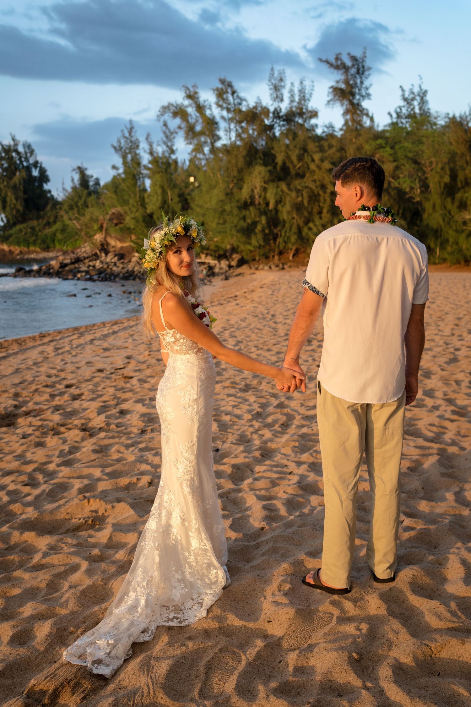 A couple walks together on a scenic beach with waves and trees in the background at sunset.