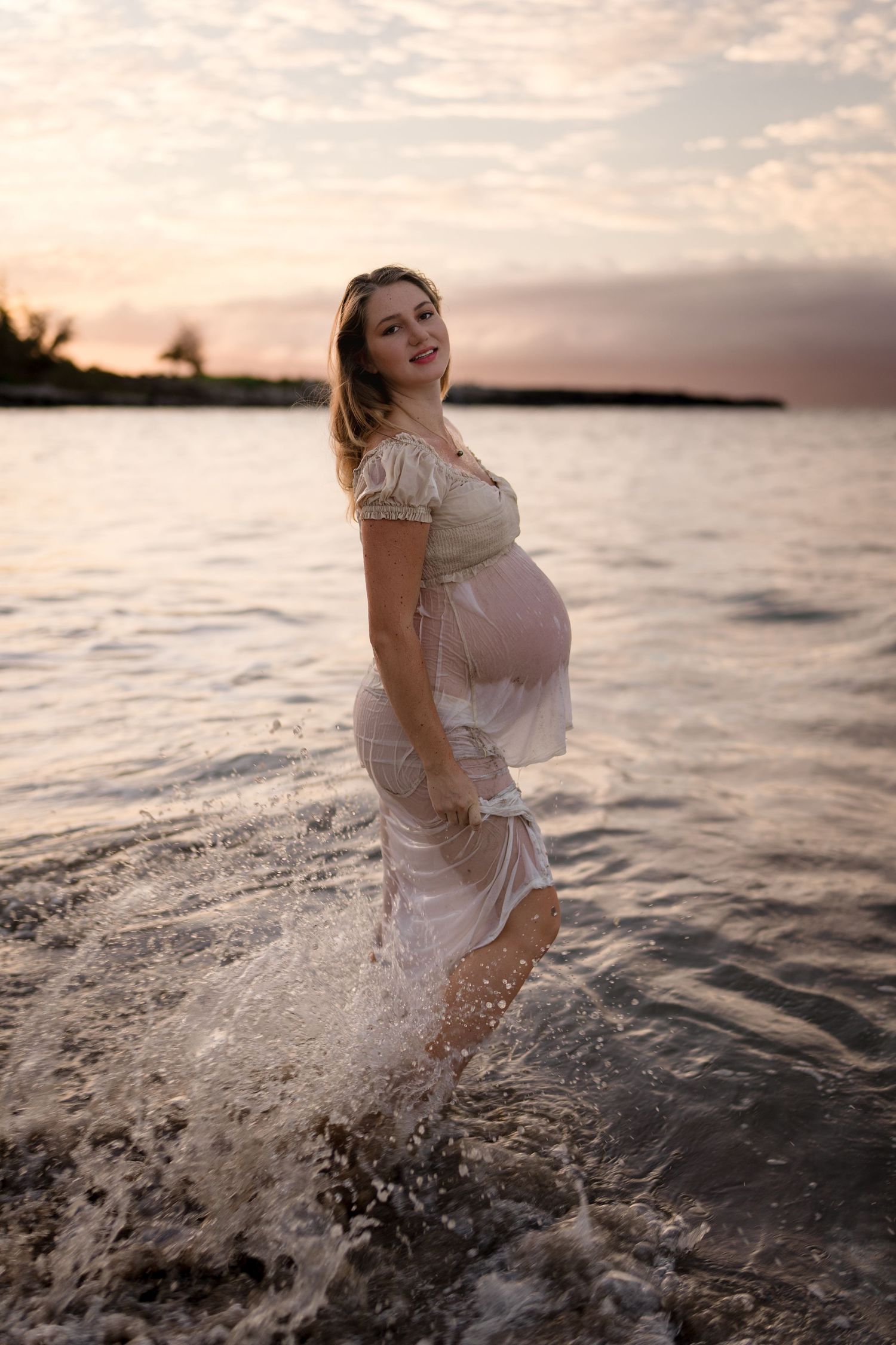 An expectant mother in a white dress splashing in ocean waves during sunset.