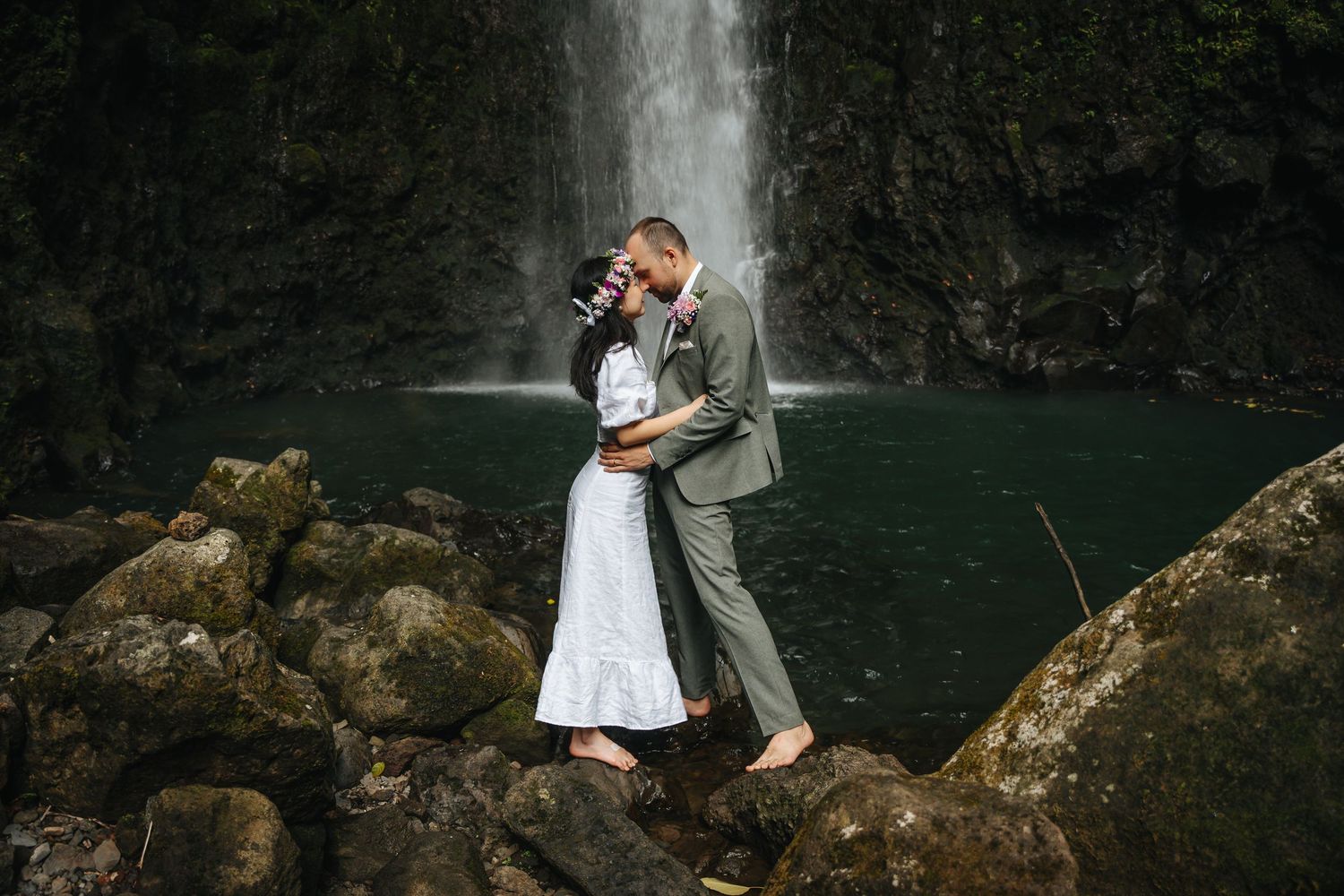 A serene waterfall cascades behind two people sitting on rocky outcrops near a pool of water.
