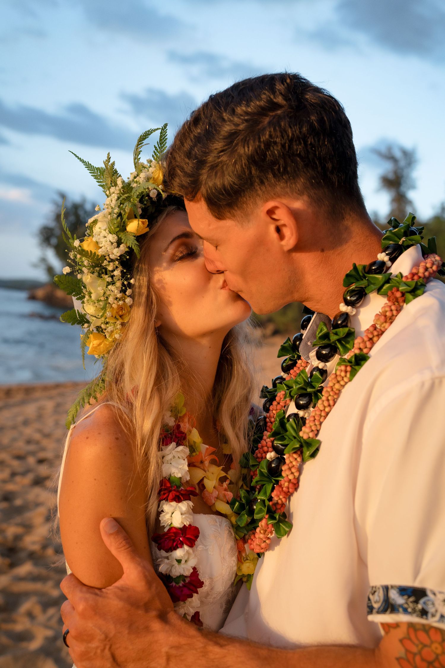A romantic beach wedding moment with floral leis and ocean backdrop at sunset.