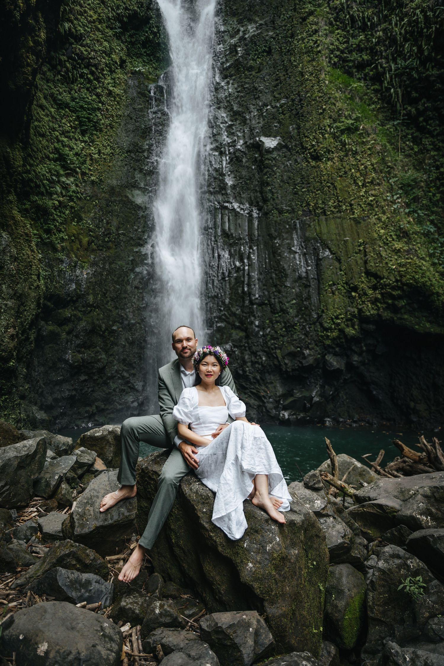A dramatic waterfall creates a stunning backdrop for a couple's portrait on rocky terrain.