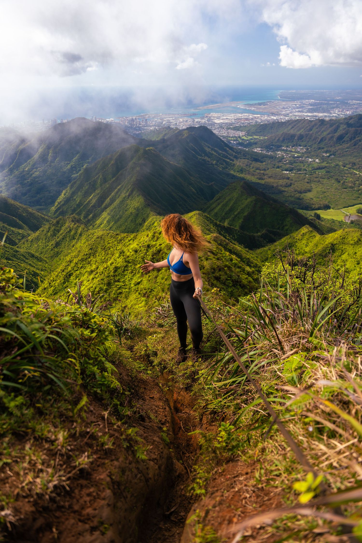 A hiker stands on a mountain ridge overlooking a vast green valley and city below.