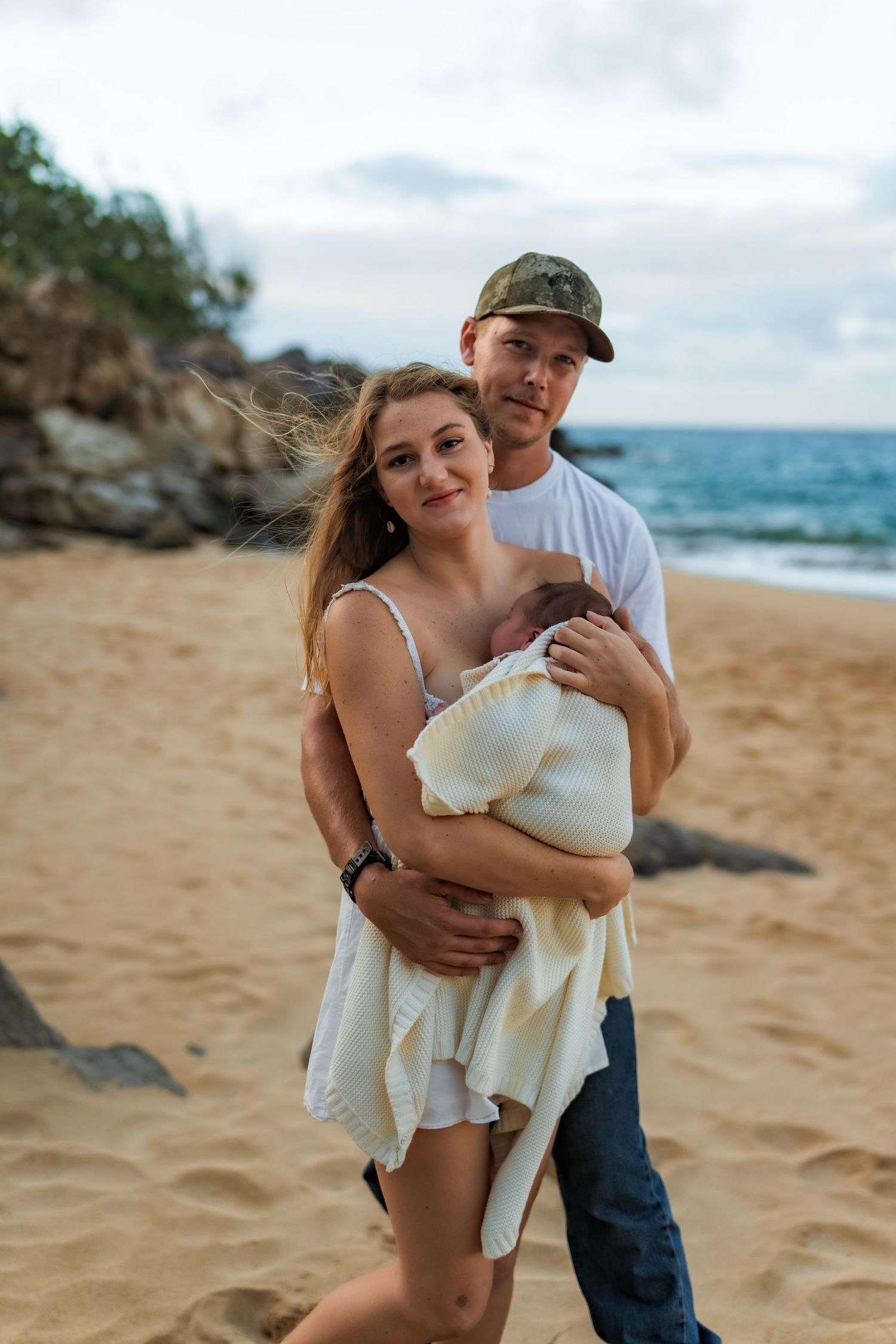 Couple sharing intimate moments with newborn on a sandy beach with ocean waves and rocks in the background.