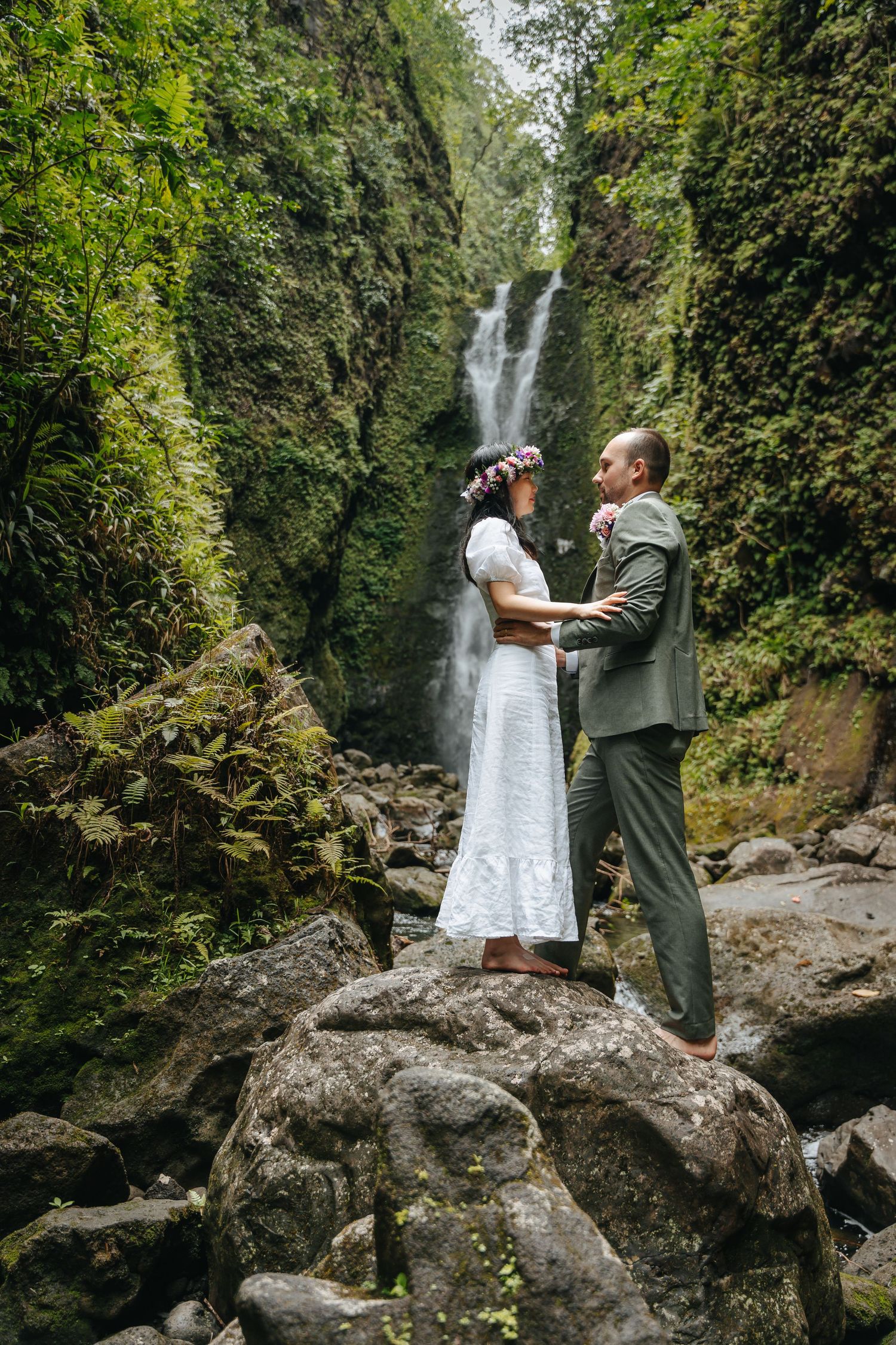 A couple shares a tender moment near a cascading waterfall in a lush forest setting.