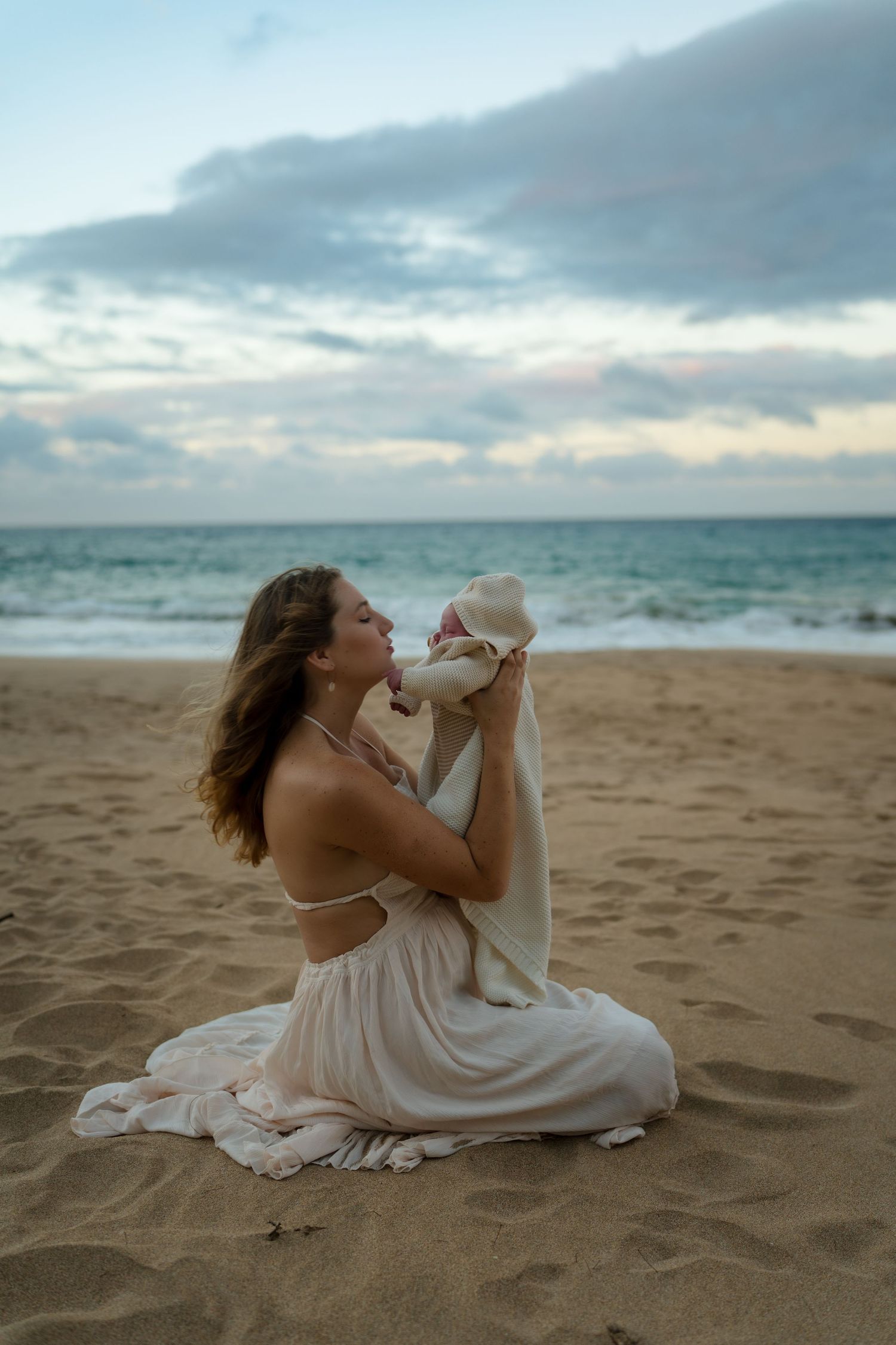 A mom in a white dress sits on a sandy beach at sunset holding her baby in a white blanket.
