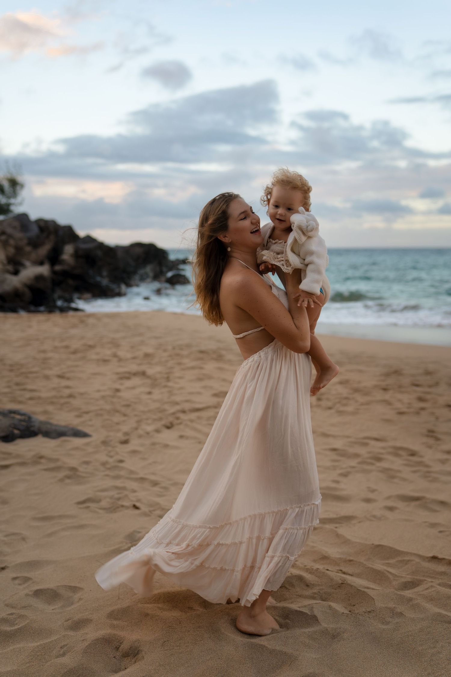 Mother in white dress plays with baby on sandy beach during golden hour.