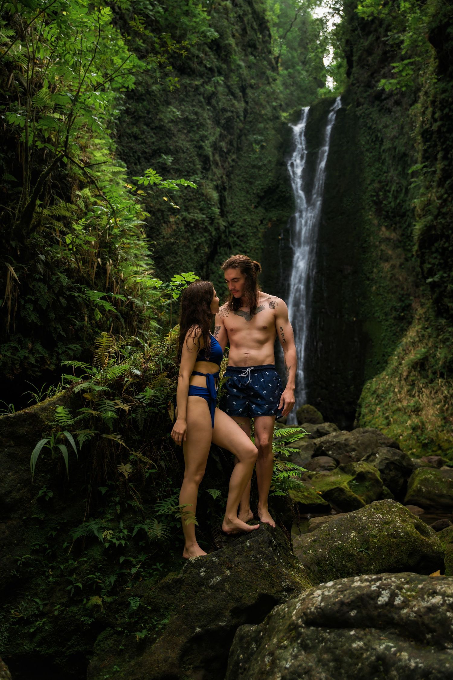 Couple relaxes on mossy rocks near a tropical waterfall in Hawaii.