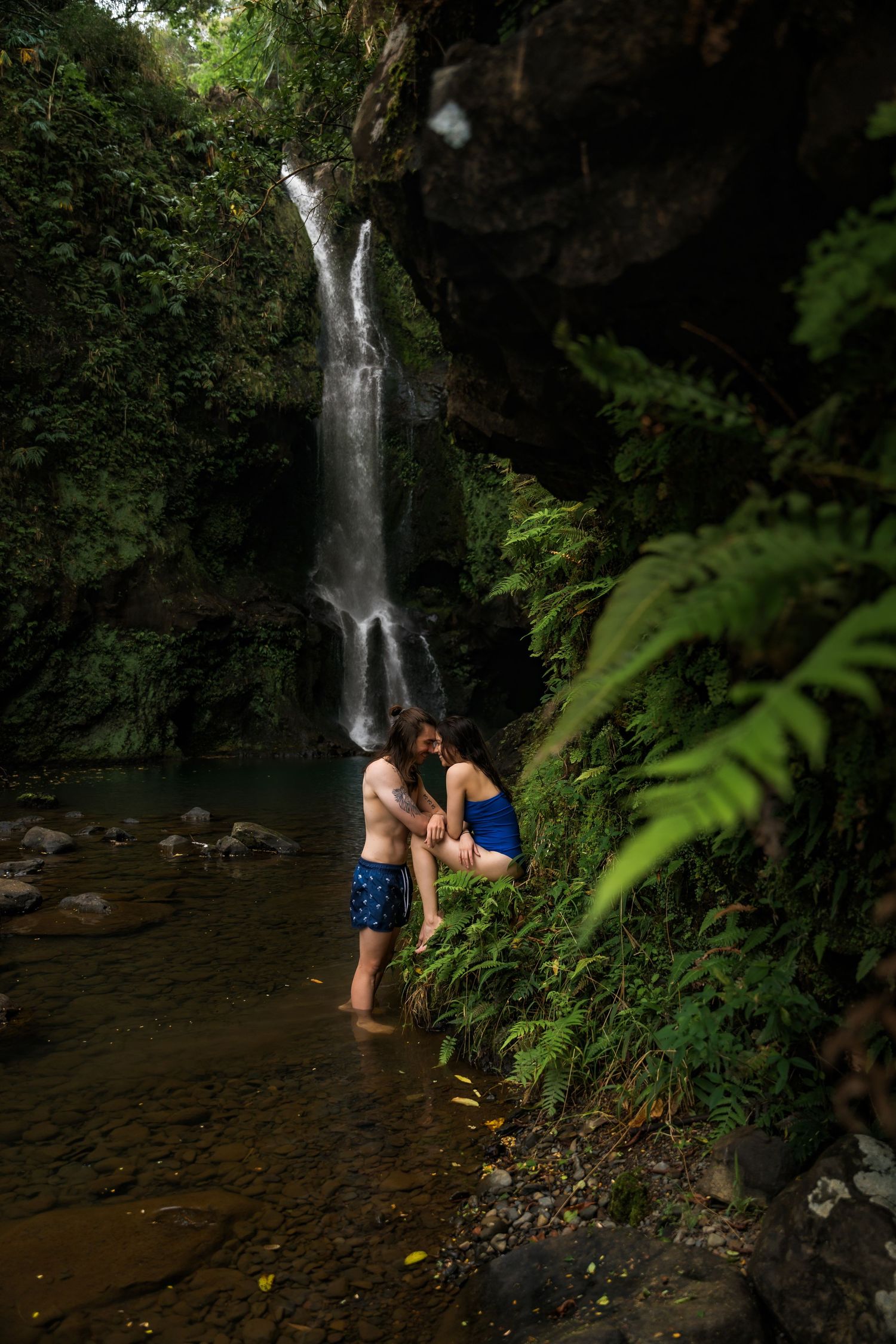 Couple embraces near a scenic waterfall surrounded by lush tropical foliage and a tranquil pool.