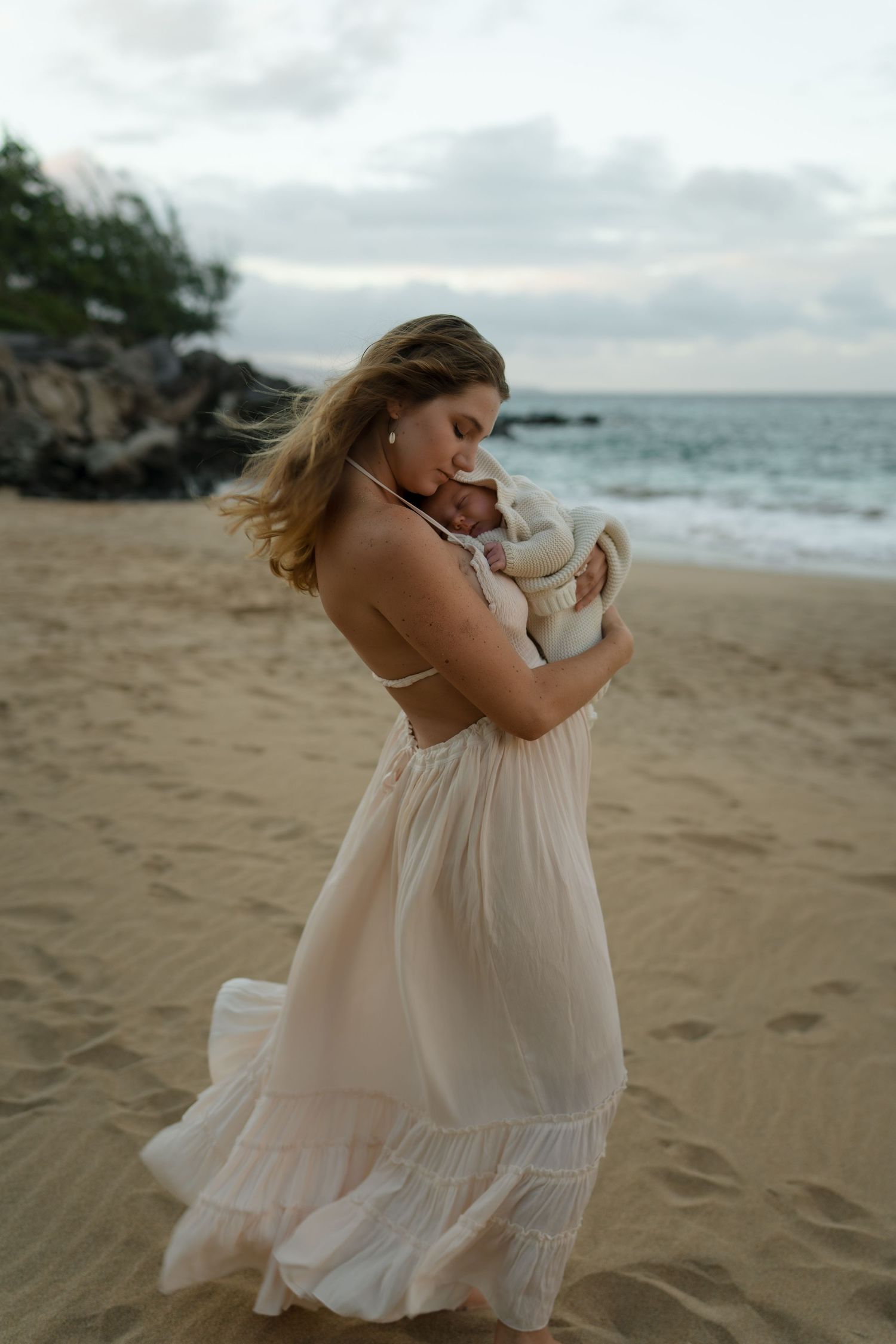 Mother in white dress embraces baby tenderly on sandy beach during golden hour.