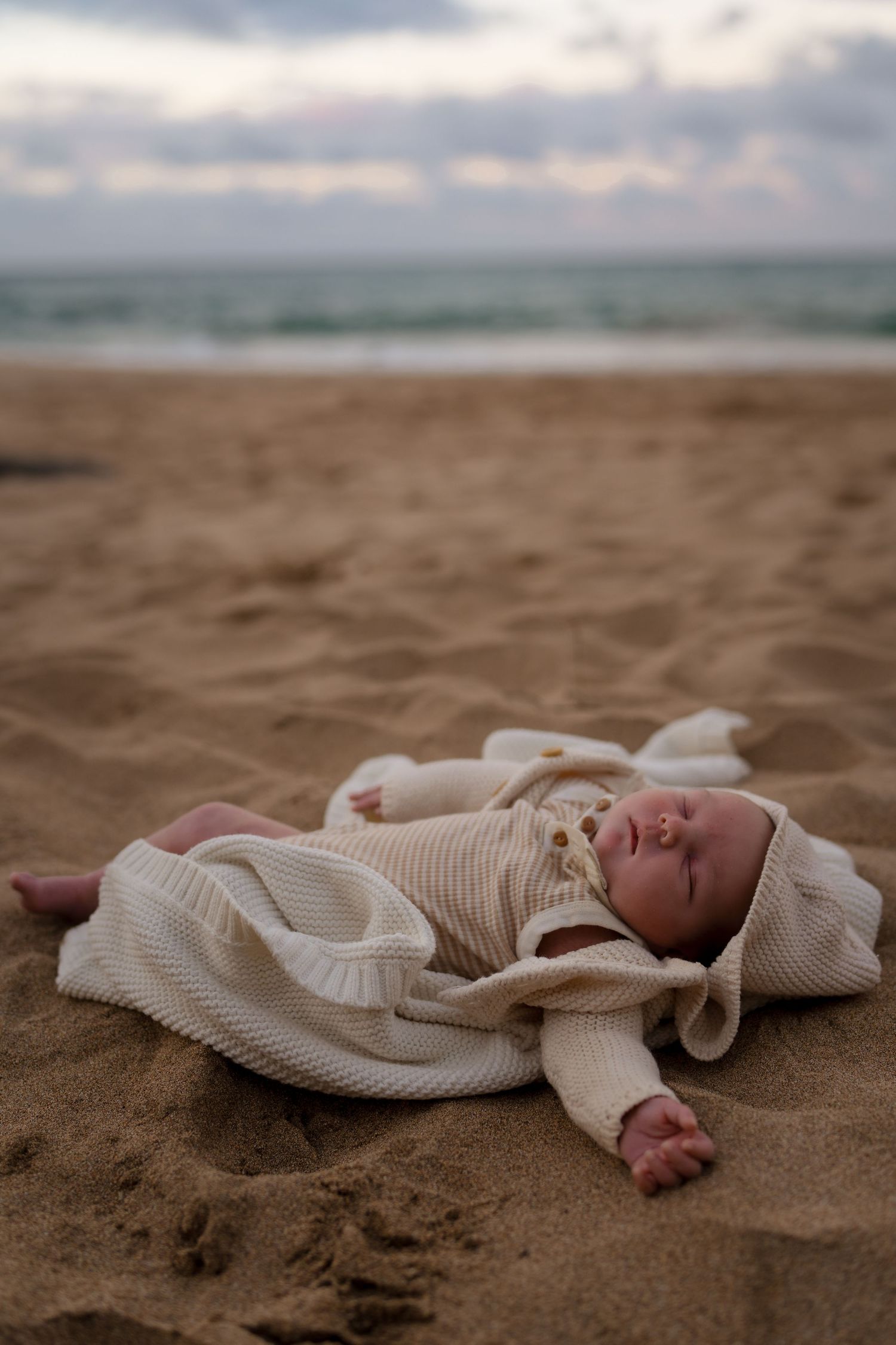 A sleeping baby in cream knit clothing lies peacefully on sandy beach.