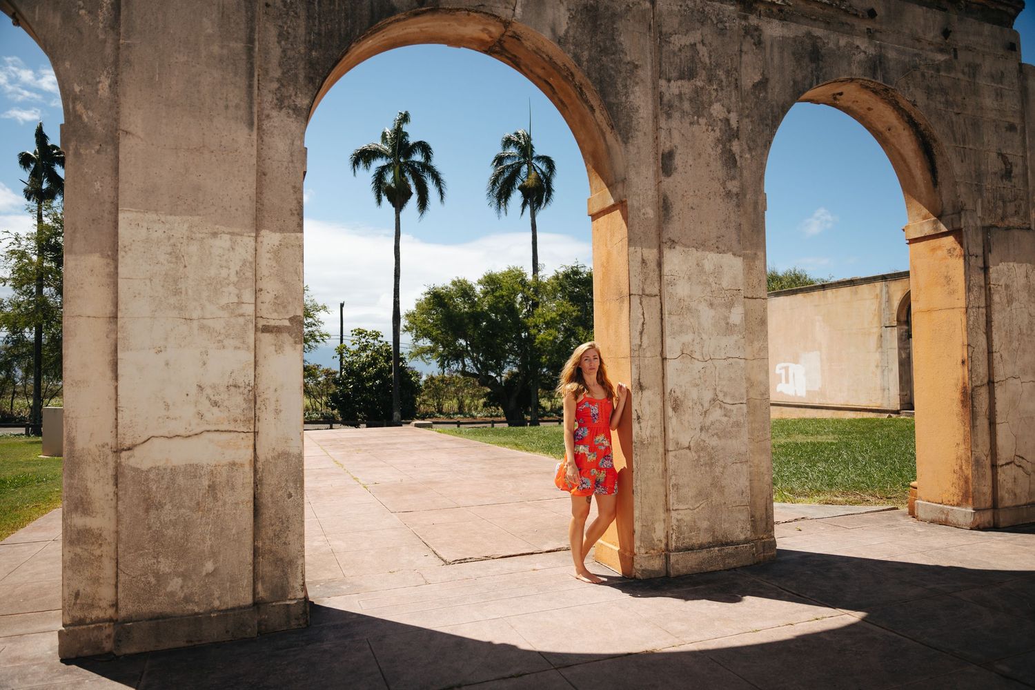 A figure in a red dress stands beneath stone archways with palm trees visible in the background.