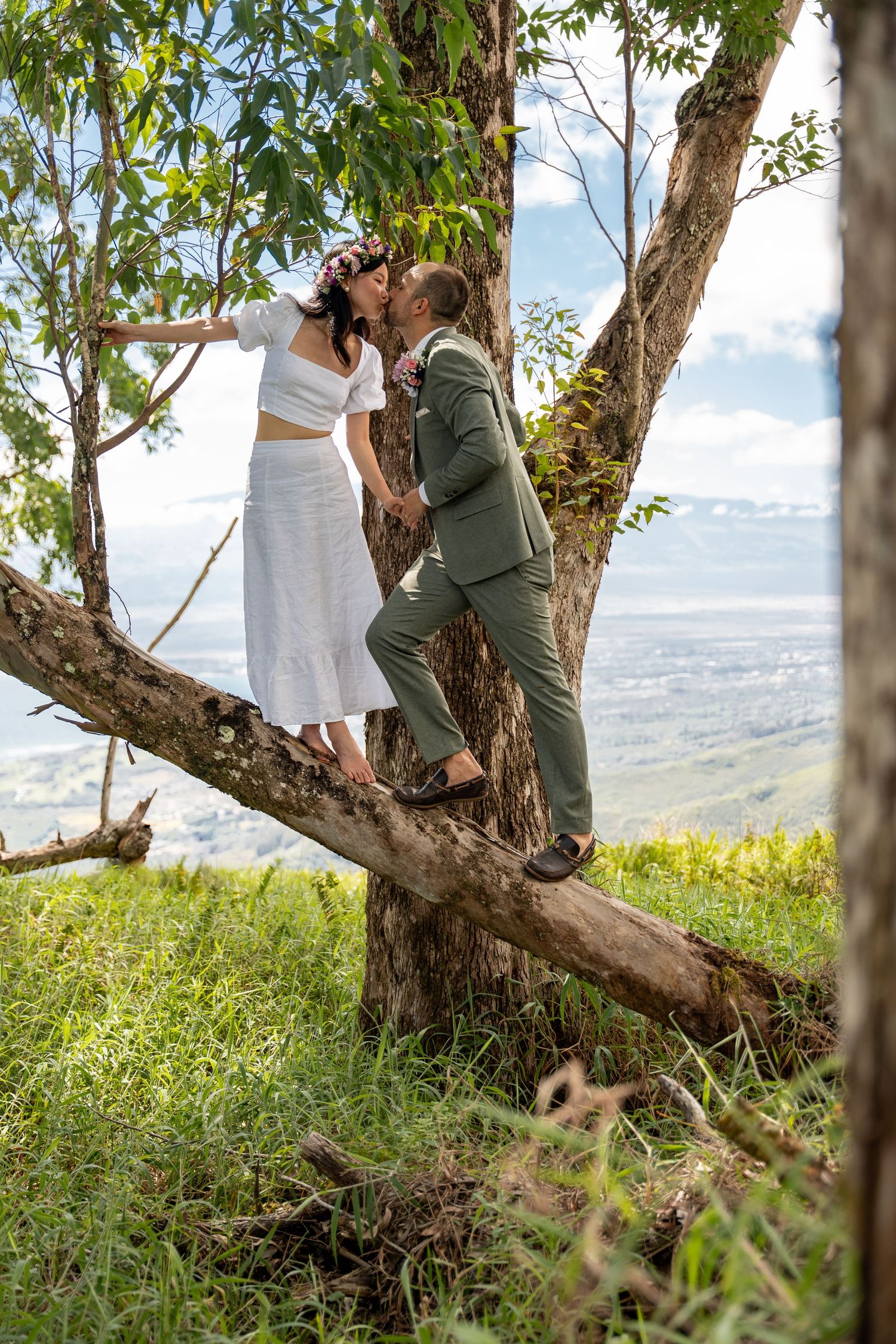 A couple in wedding attire poses playfully on a tree branch overlooking a scenic mountain landscape.