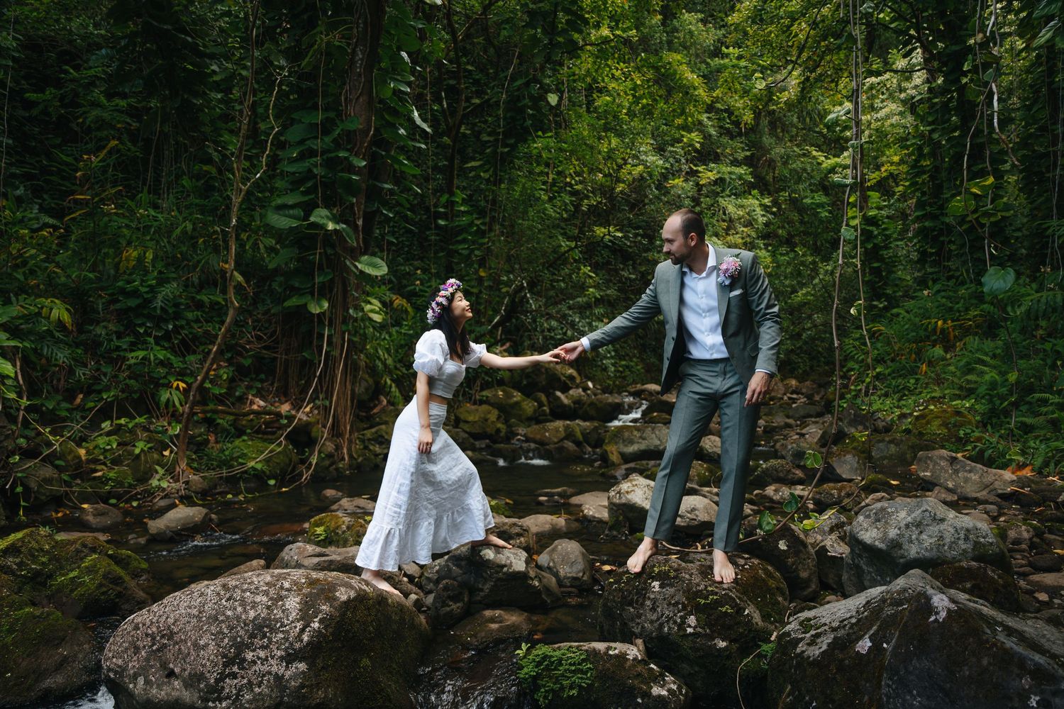 A couple explores a forest path surrounded by moss-covered rocks and dense vegetation.