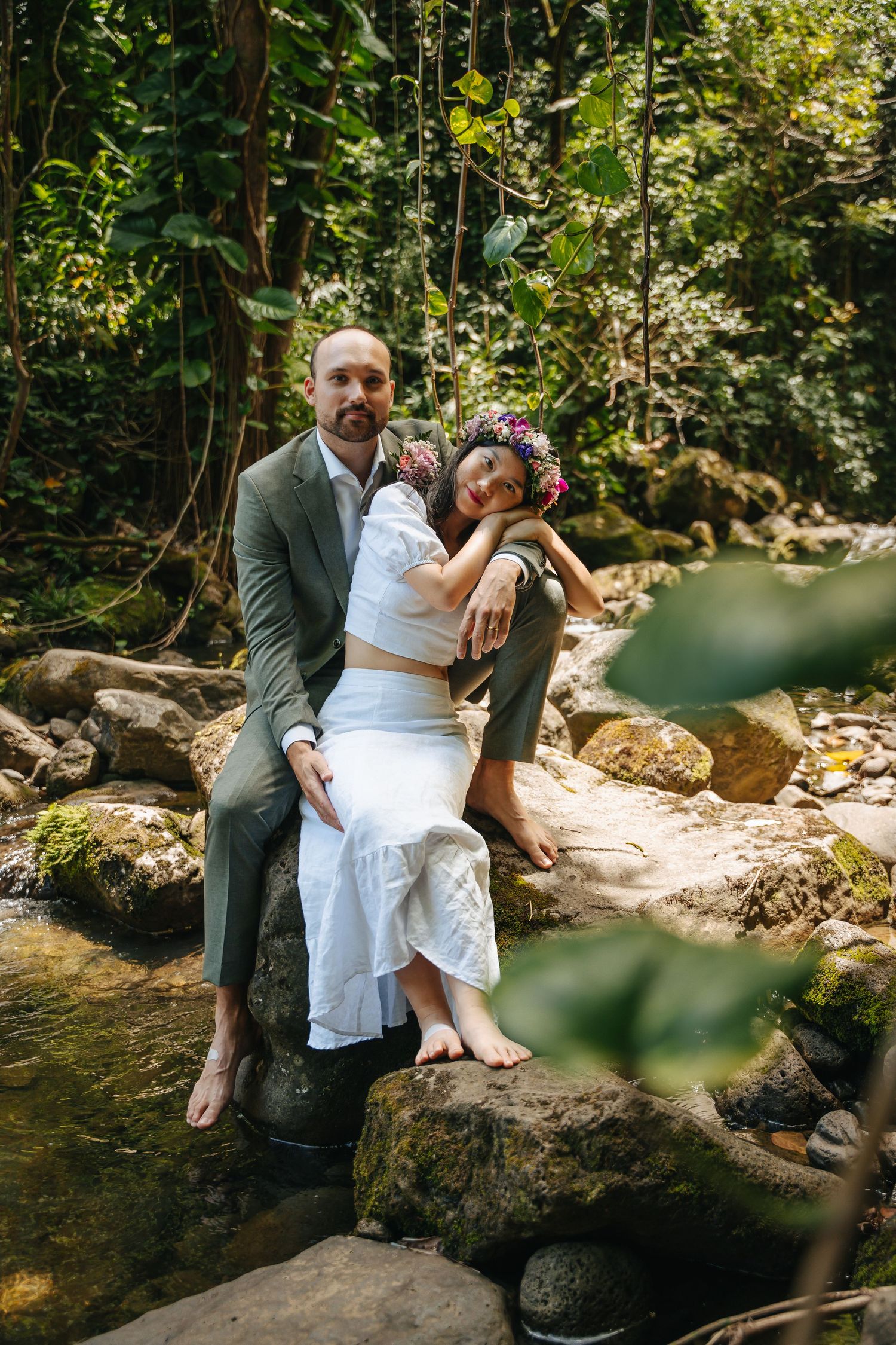 Two people share an intimate moment while sitting on rocks beside a flowing stream in a forest.