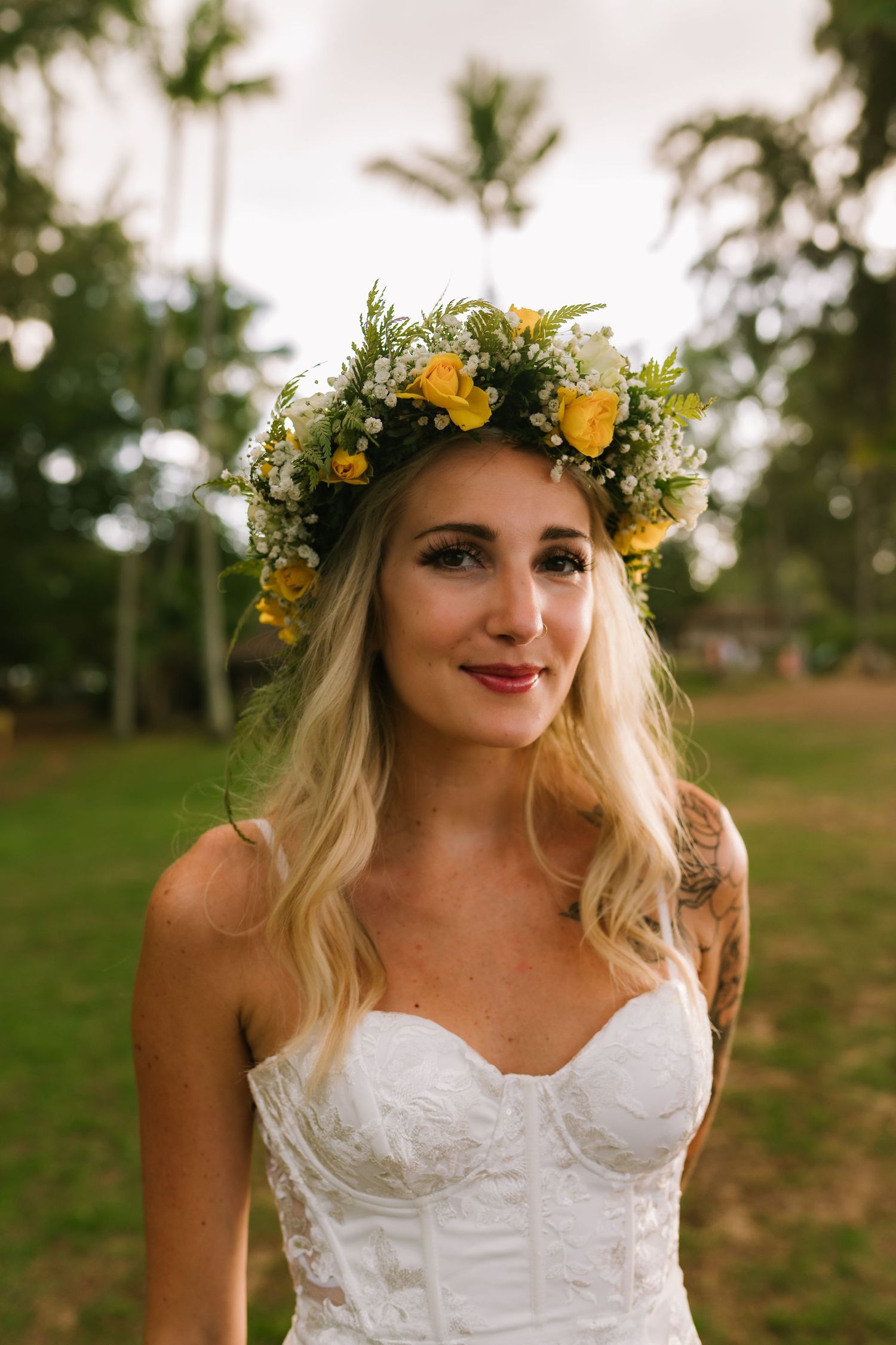 A person wearing a yellow flower crown and white dress smiles in warm outdoor lighting.