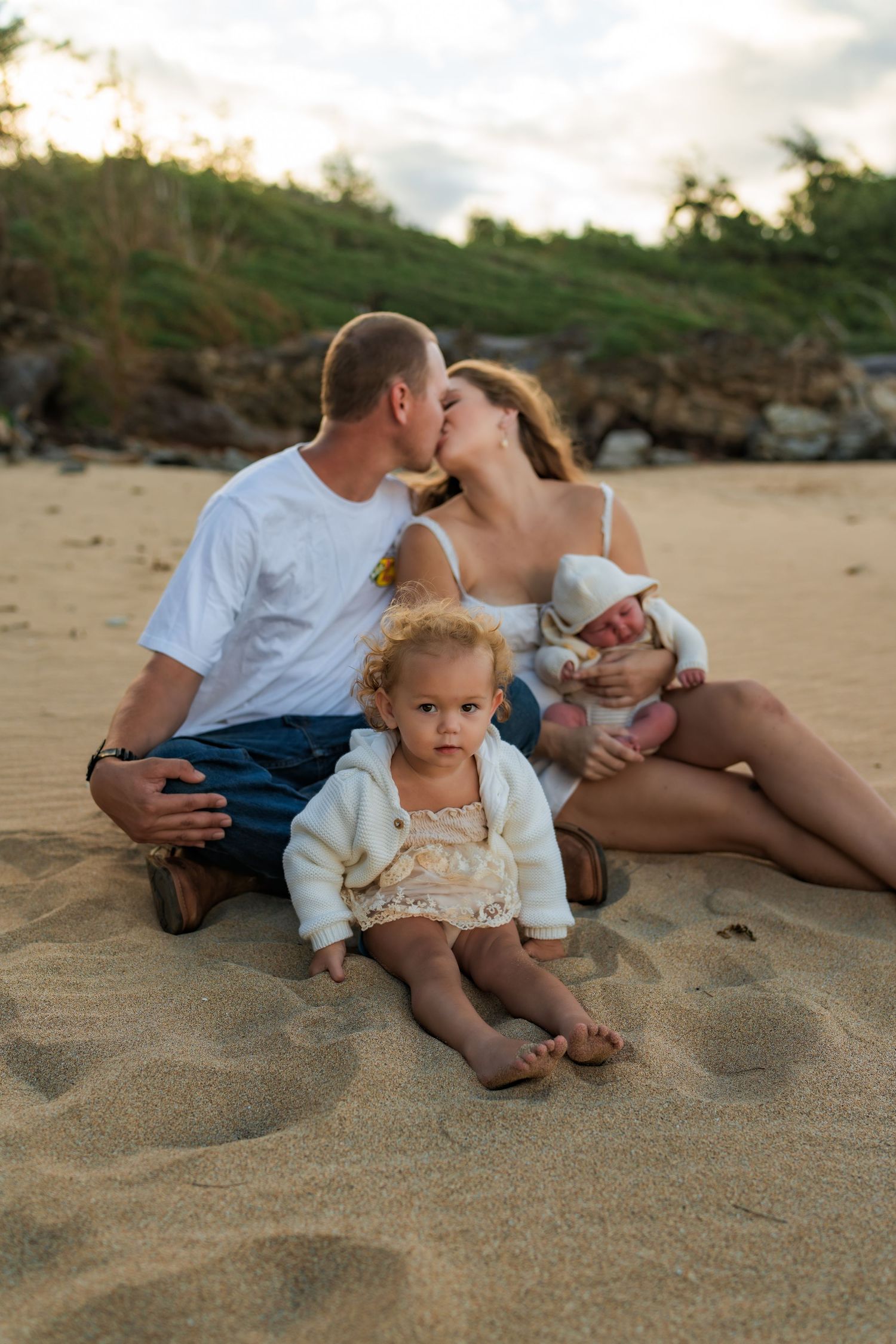 A family relaxes together on the sandy beach at sunset with rocky cliffs in the background.