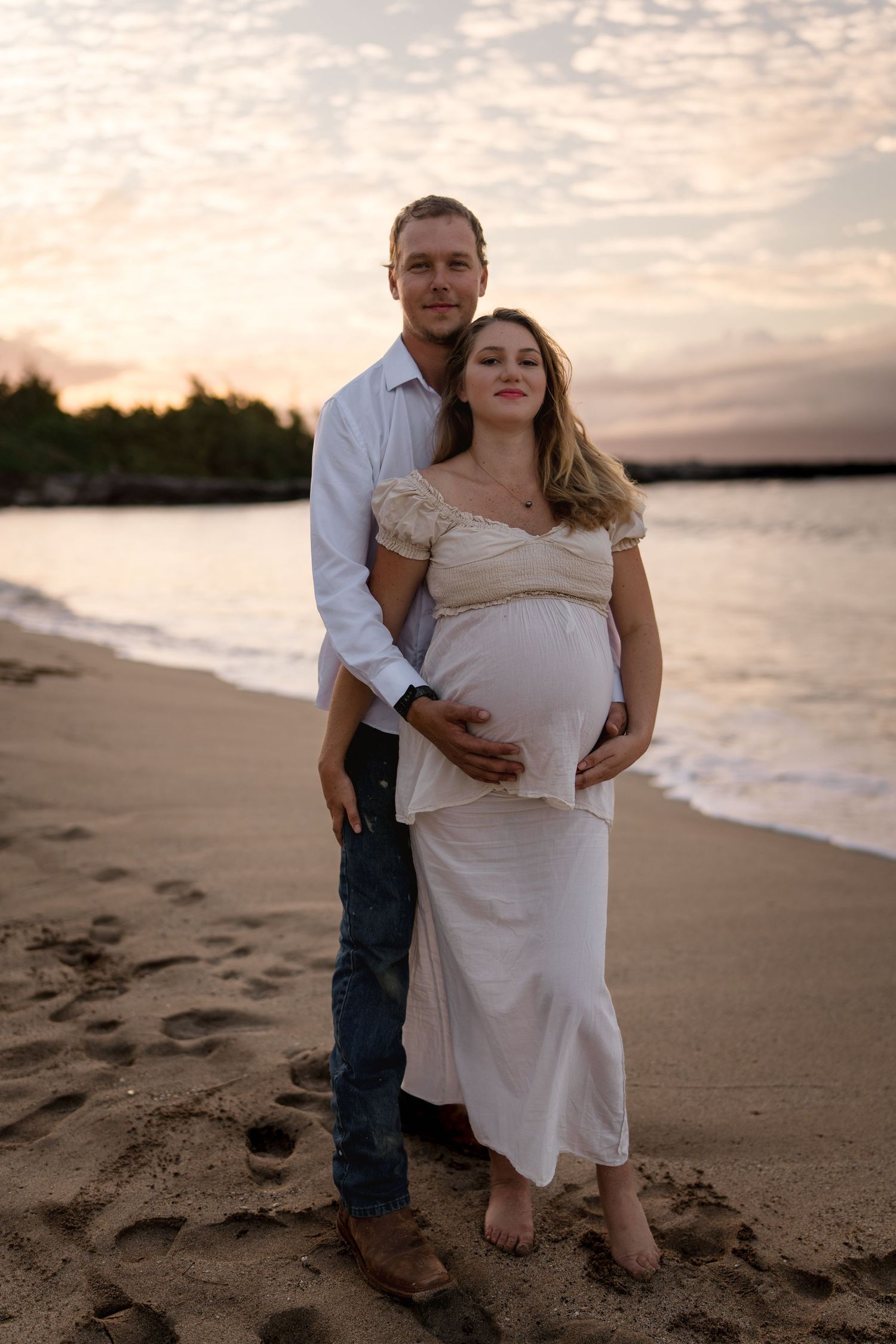 Intimate maternity photoshoot on beach at sunset with couple in white and denim.