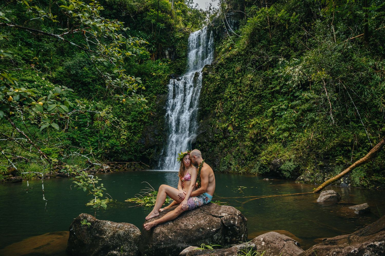 Multiple waterfalls cascade down rocky cliffs into a tranquil swimming hole in a tropical forest setting.