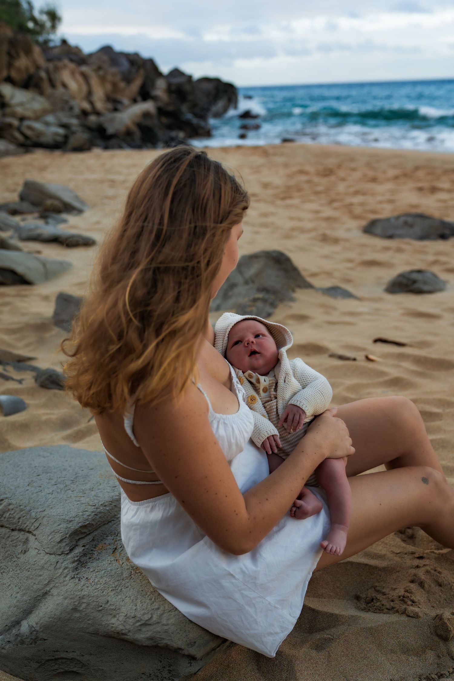A mom in a white dress holds her baby while sitting on a rocky beach with ocean waves in the background.