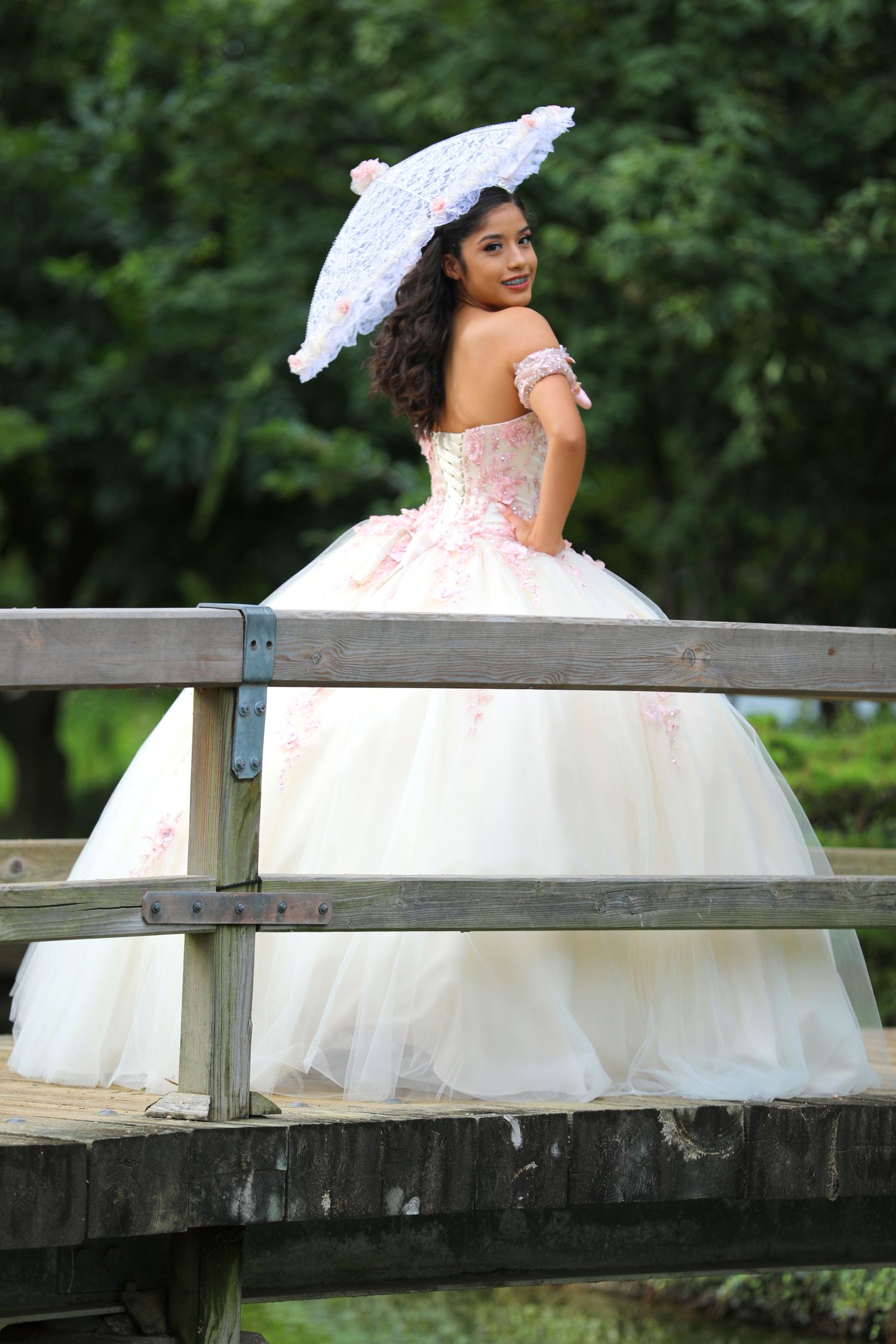A person in an elegant pink quinceañera dress poses with a white parasol on a wooden bridge.