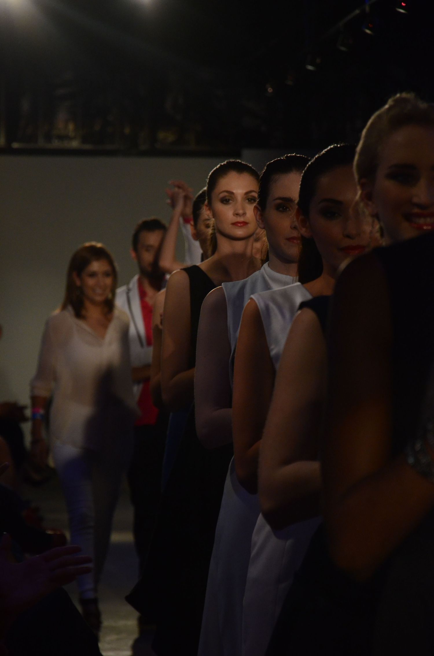 Models lined up backstage at fashion show in dimly lit setting.