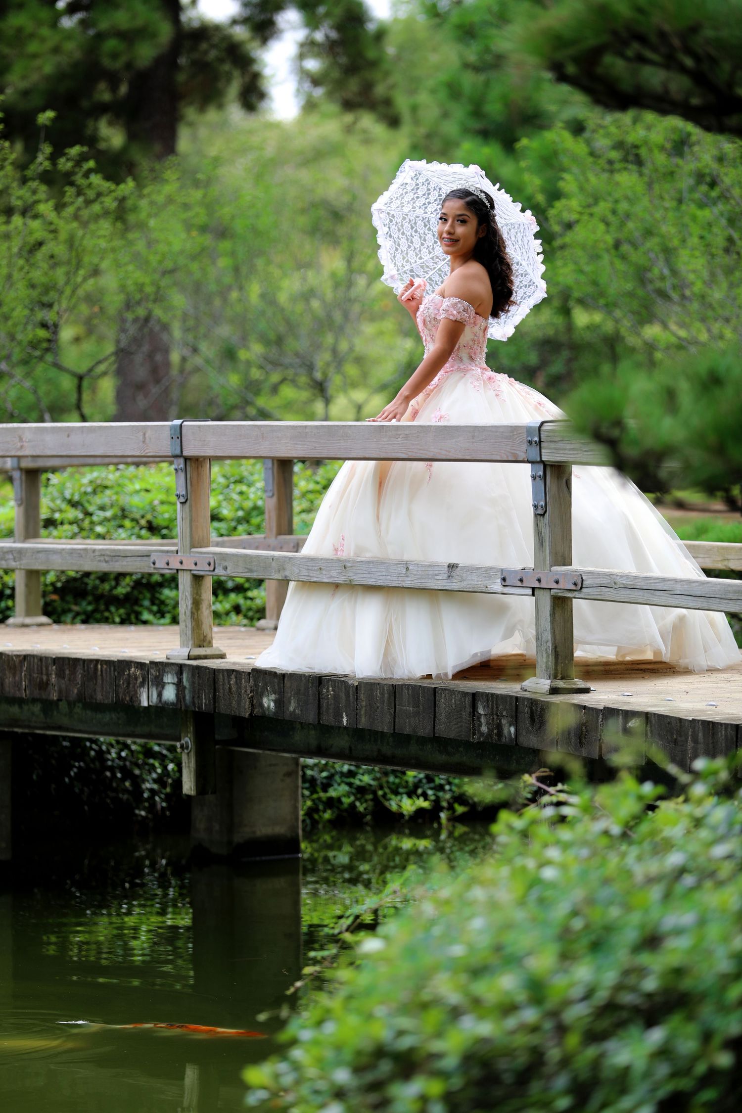 A person in a white gown with a lace parasol poses on a wooden bridge in a garden setting.