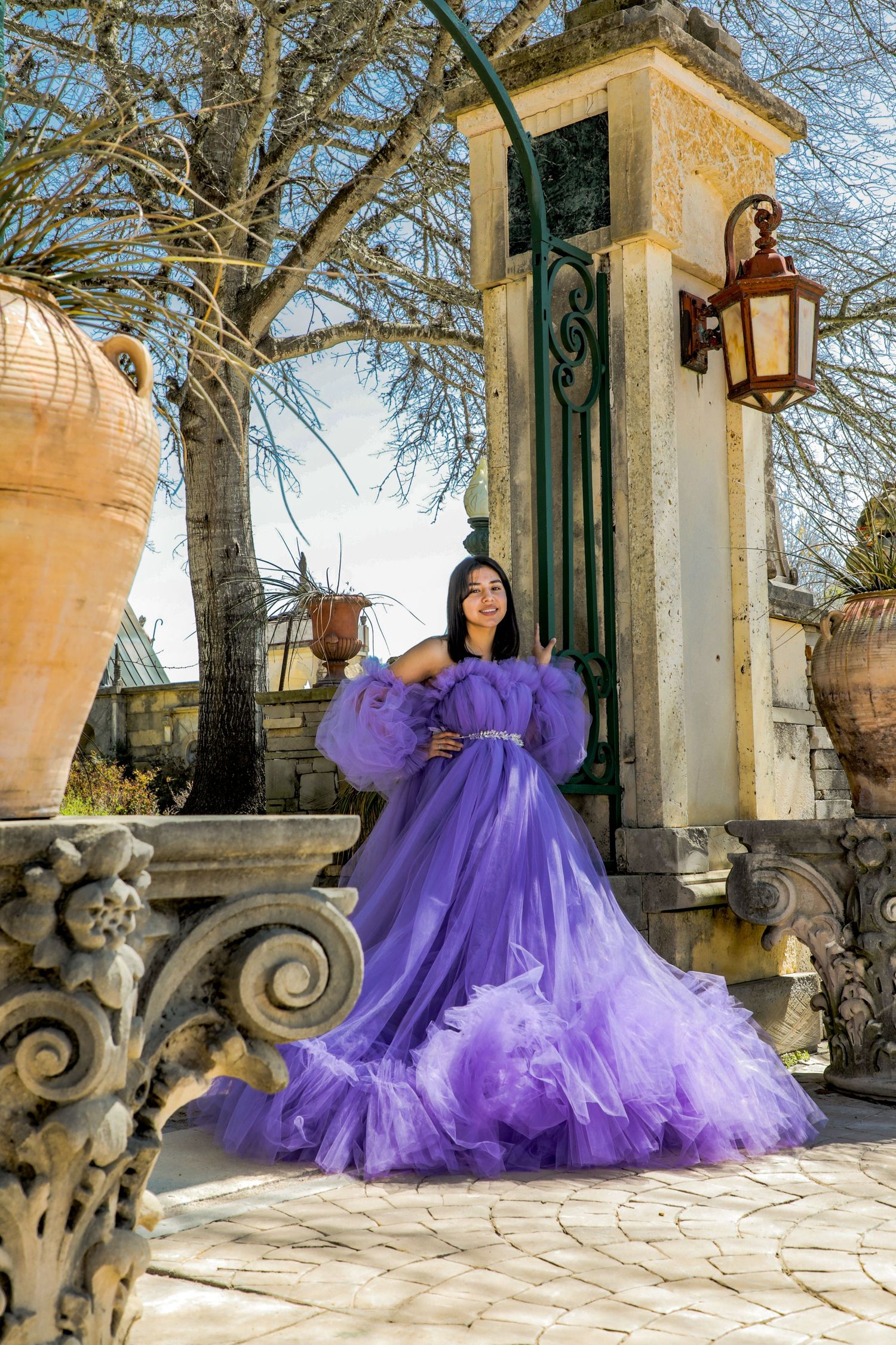 A dramatic purple tulle gown with billowing sleeves photographed against a stone pillar and lantern in an elegant outdoor setting.
