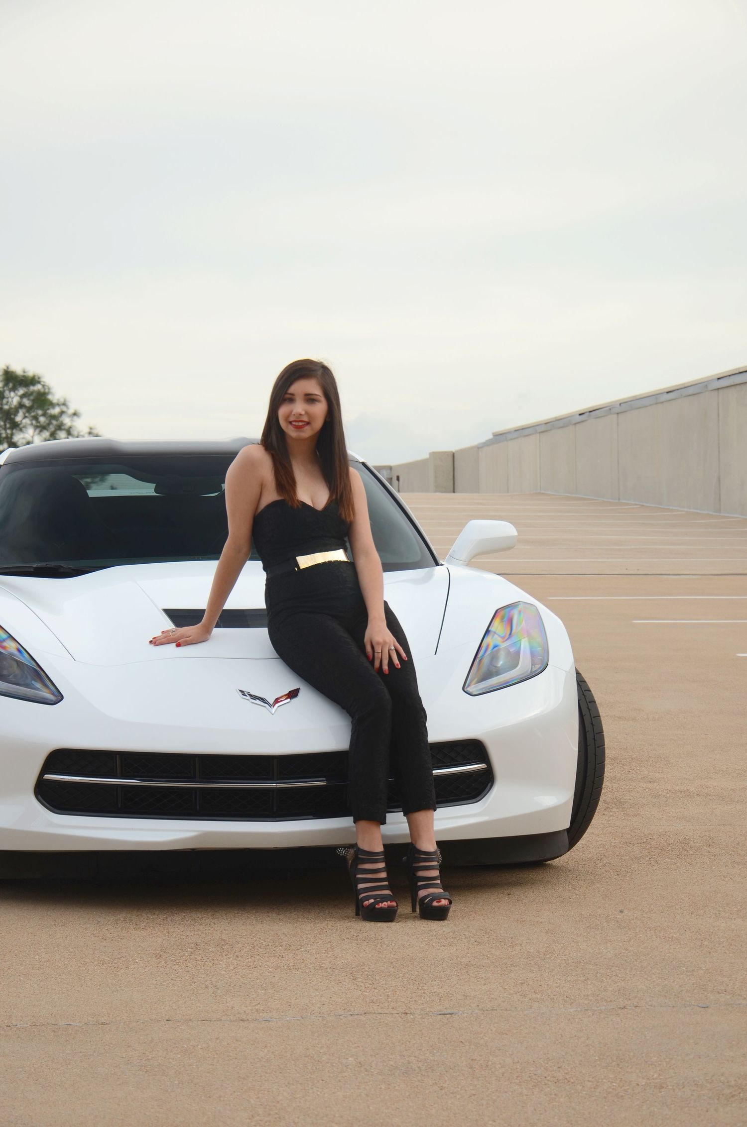 White Corvette sports car with figure in black outfit posed on hood.