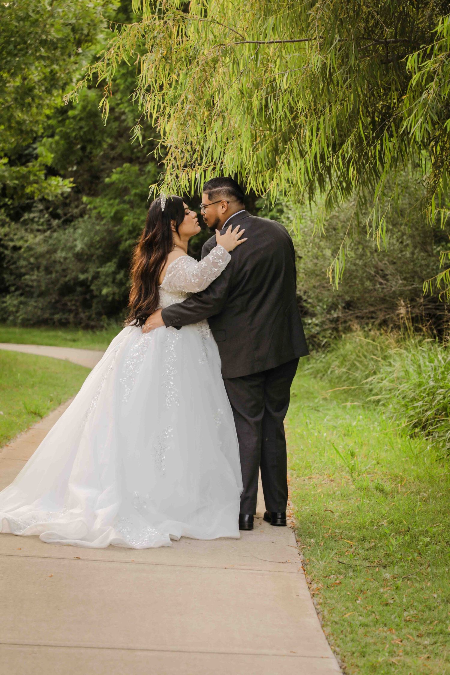 A wedding couple shares a romantic kiss underneath a weeping willow tree on a garden path.